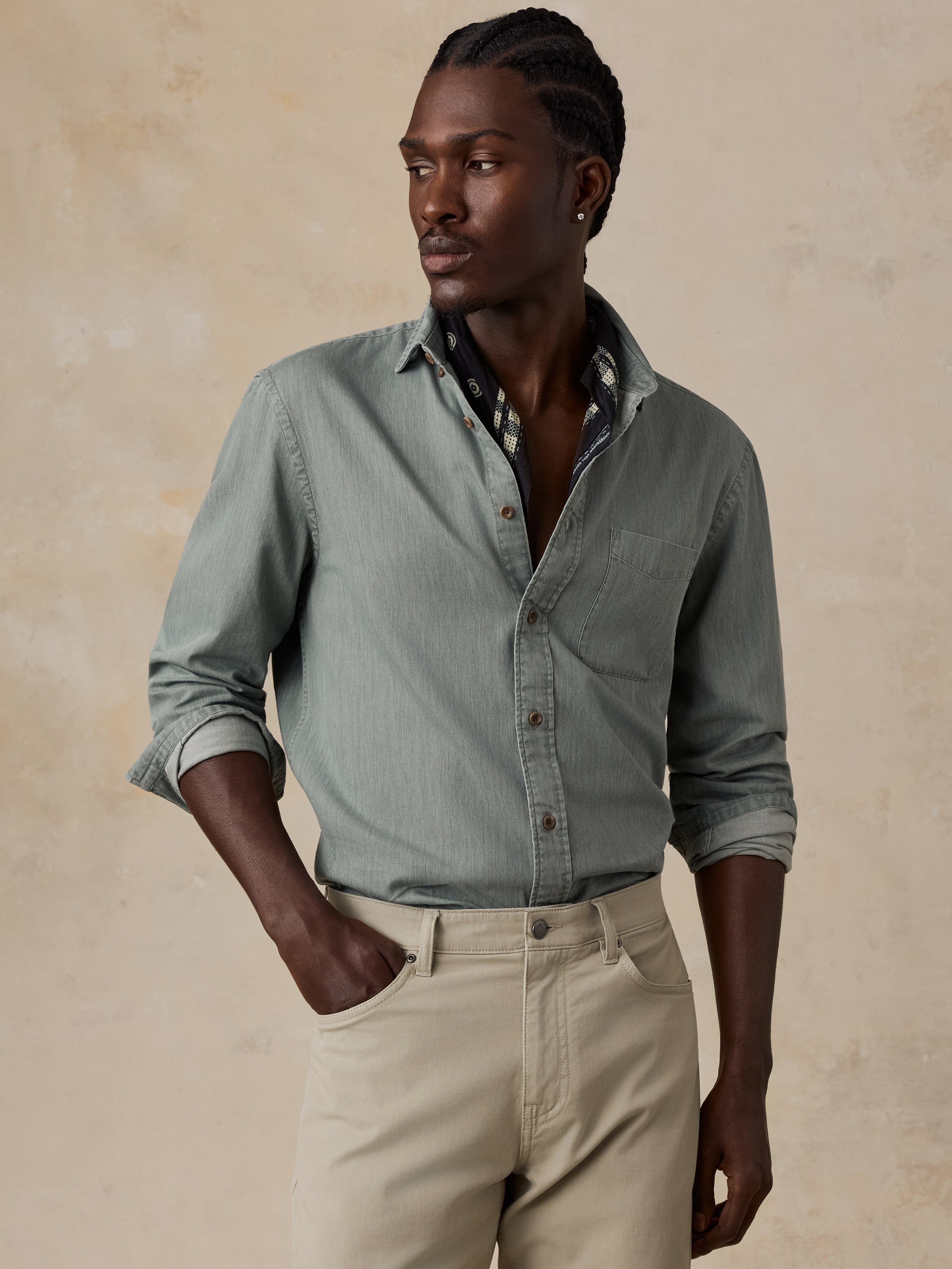 A man wearing a sage green long-sleeve button-down shirt and beige trousers, standing against a textured beige background.