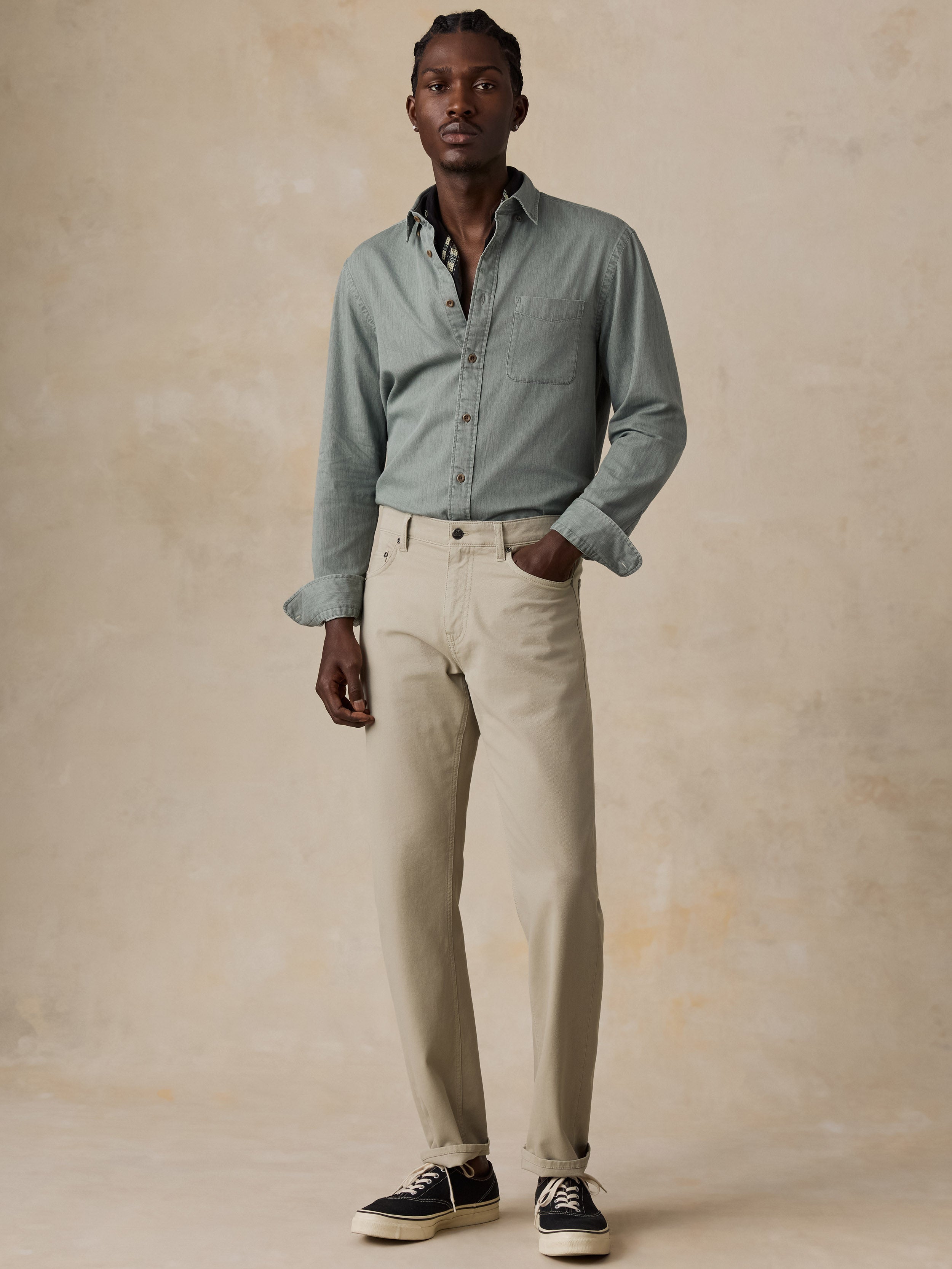 A man modeling a light sage green long-sleeve button-down shirt and beige trousers, paired with black sneakers, against a textured beige studio backdrop.