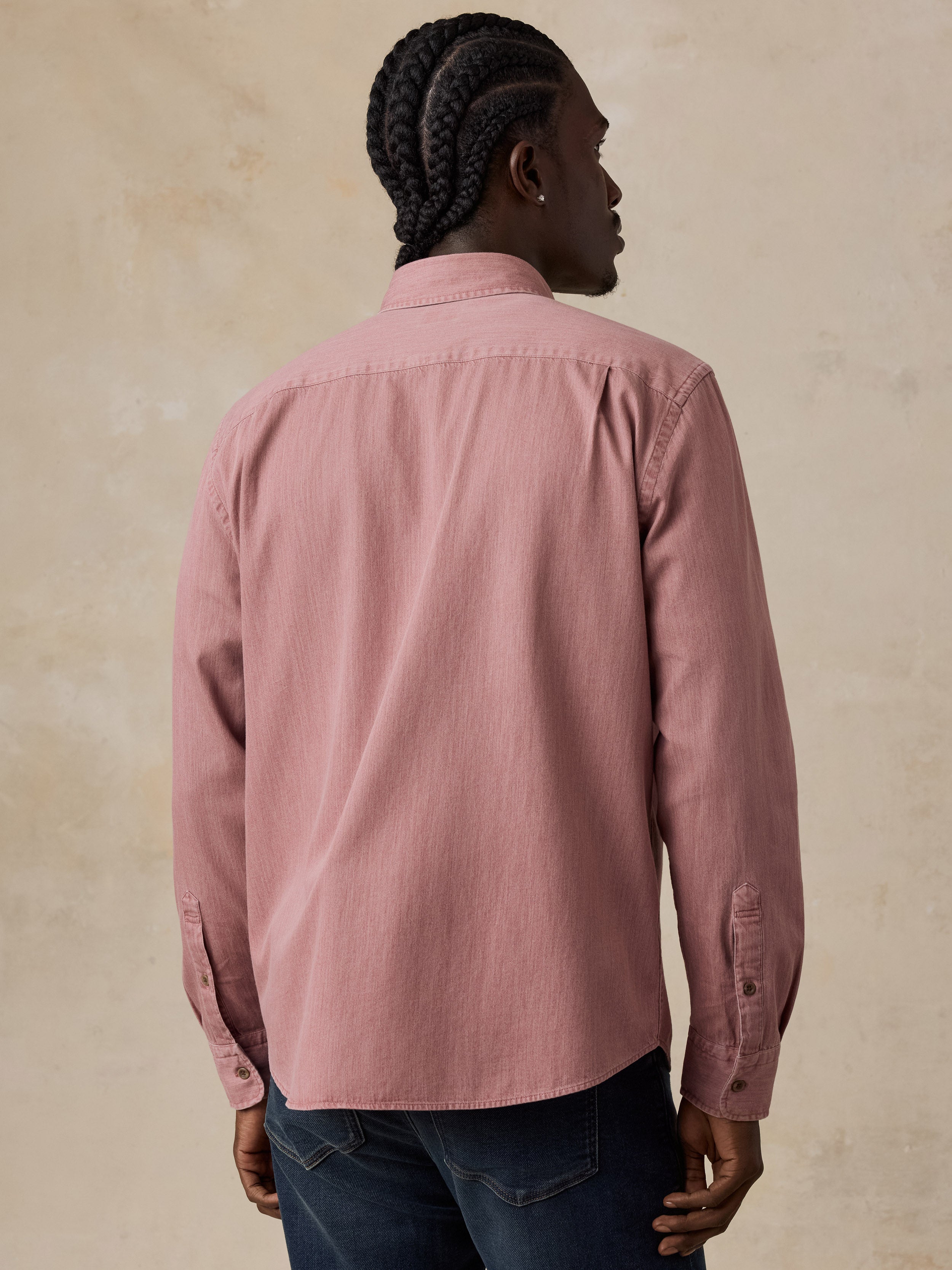 A man viewed from behind wearing a dusty rose pink long-sleeve button-down shirt and dark blue jeans, standing against a textured beige wall.