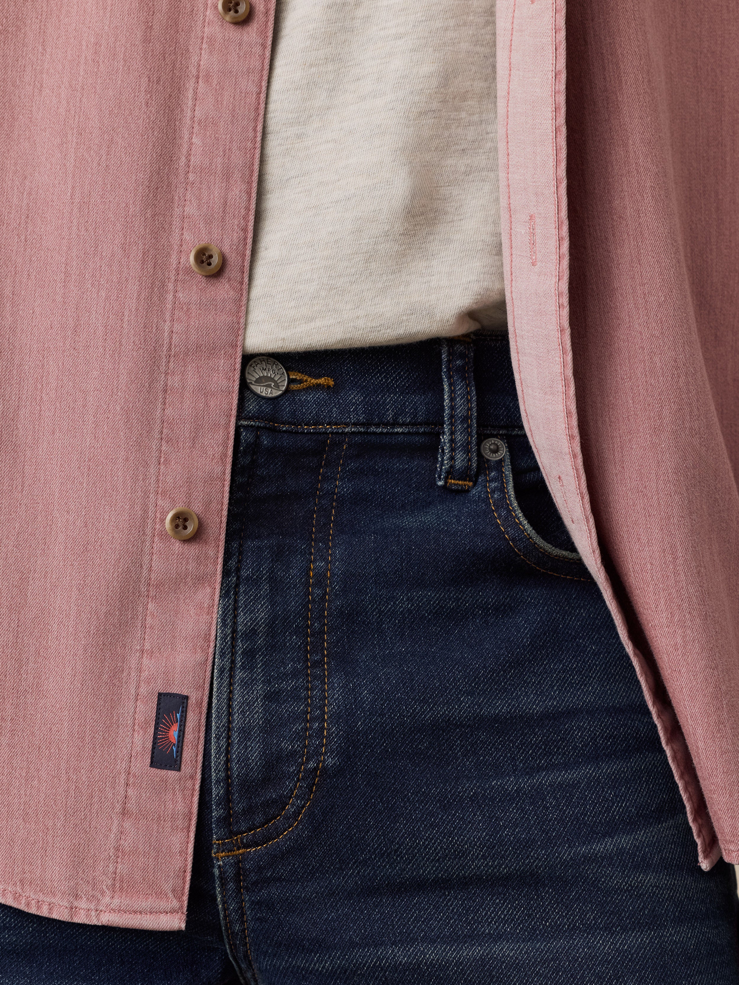 Close-up of a man wearing a dusty pink button-up shirt over a light beige tee, paired with dark blue jeans.