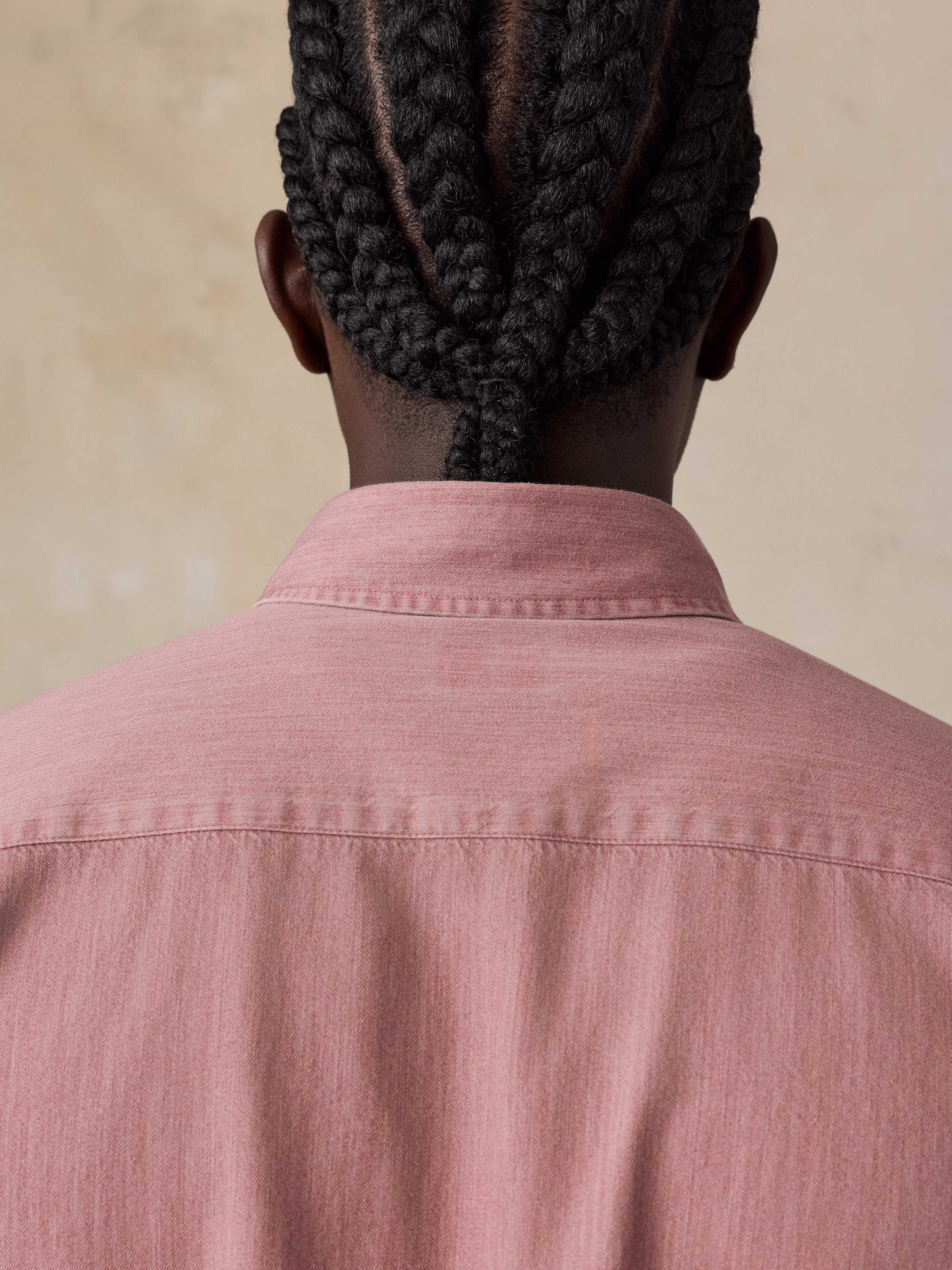 Back view of a man with dark braided hair wearing a dusty pink button-up shirt against a warm beige background.