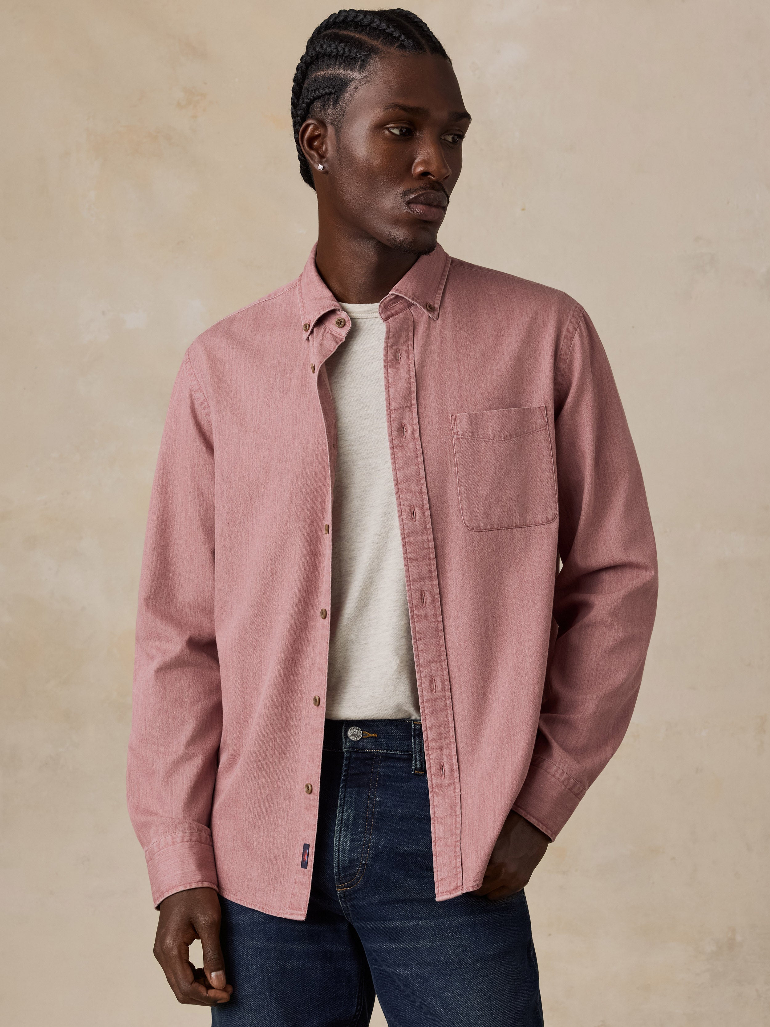A man wearing a dusty pink long-sleeve button-up shirt over a cream t-shirt and dark blue jeans, standing against a textured beige background.