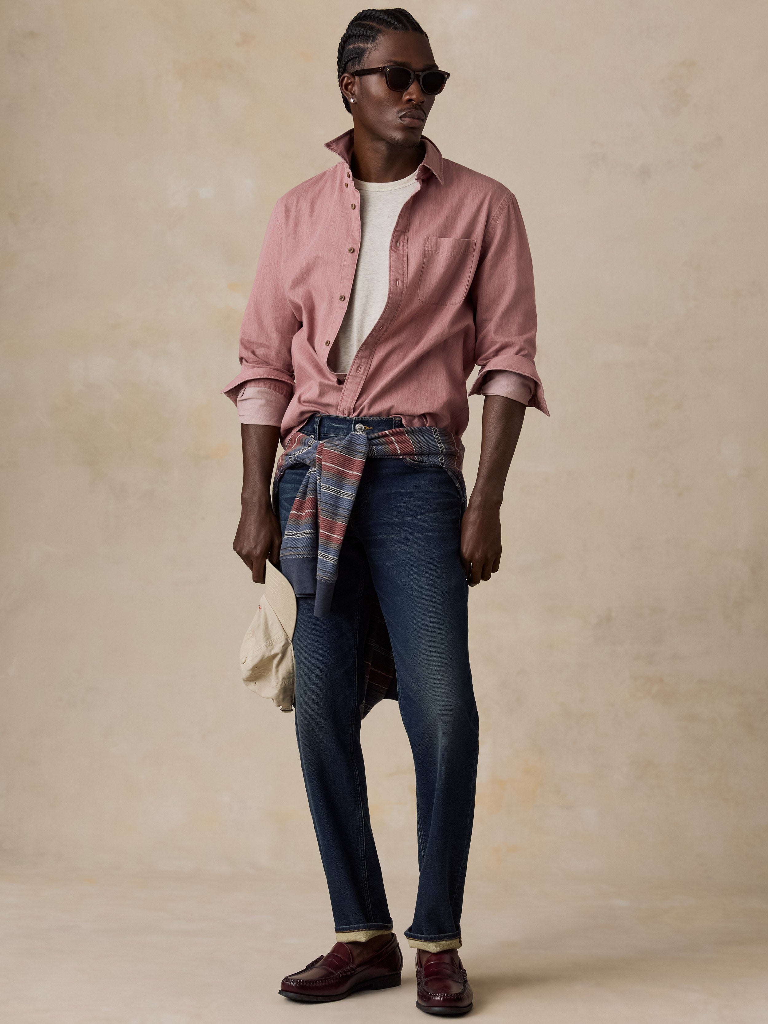 A man modeling a dusty pink button-down shirt tied at the waist over a plaid shirt, dark blue jeans, a white t-shirt, and brown loafers, set against a textured beige studio backdrop.