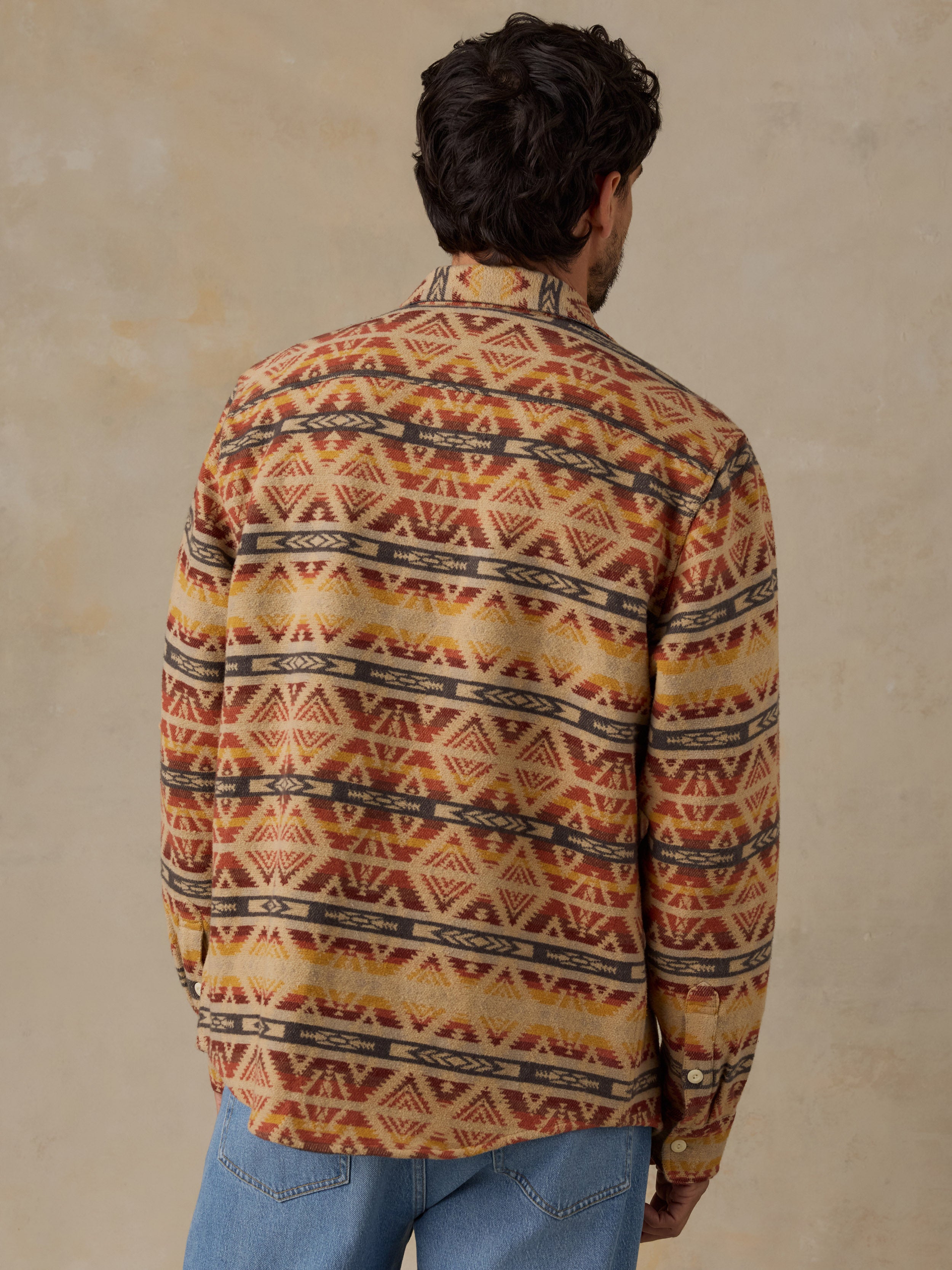 A man viewed from behind wearing a long-sleeve button-up shirt with a Southwestern geometric pattern in orange, brown, and beige, paired with blue jeans against a textured beige background.