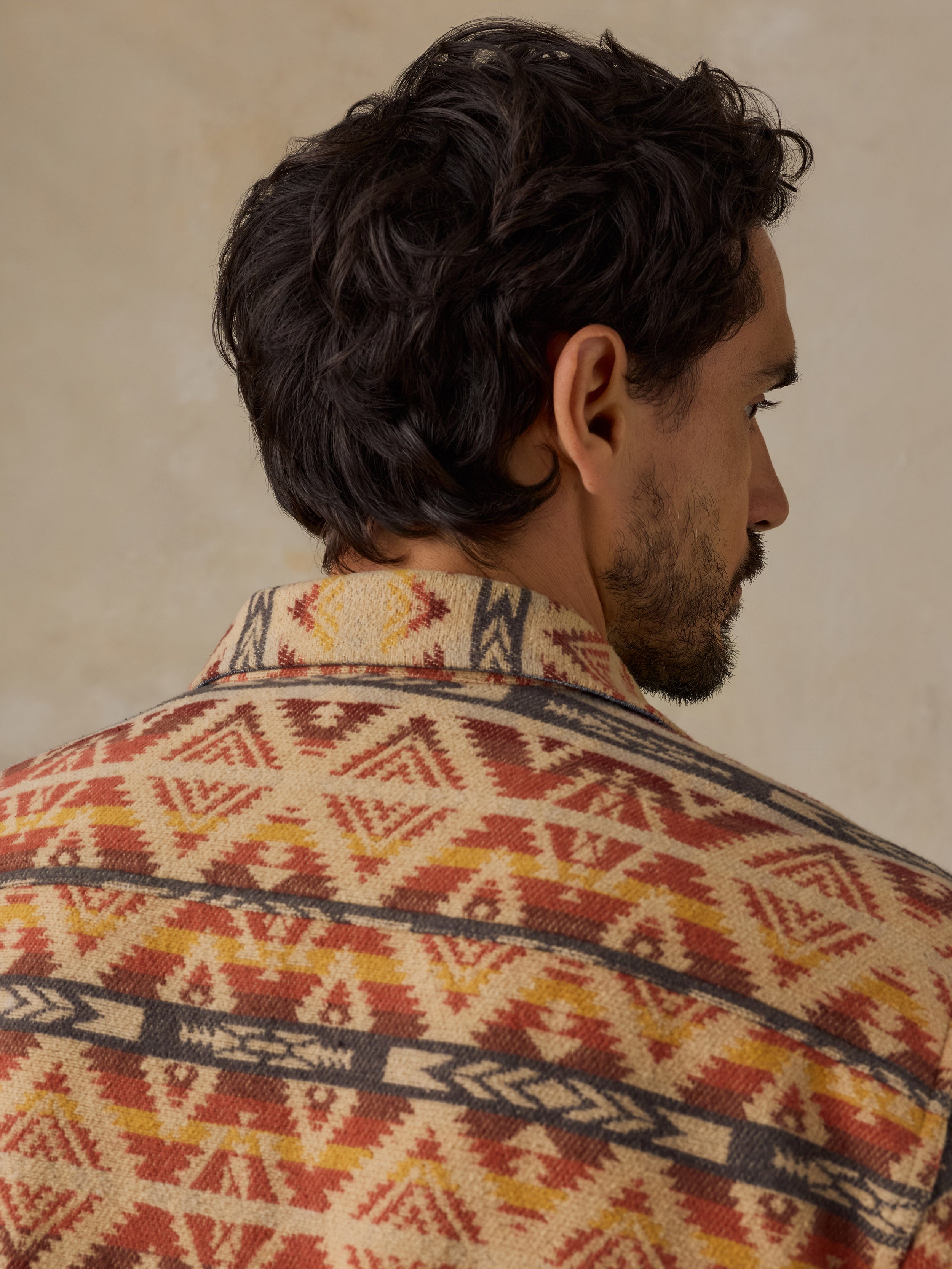 A man shown from the back and side profile wearing a patterned knit shirt with a Southwestern geometric design in red, yellow, and dark gray on a cream background.