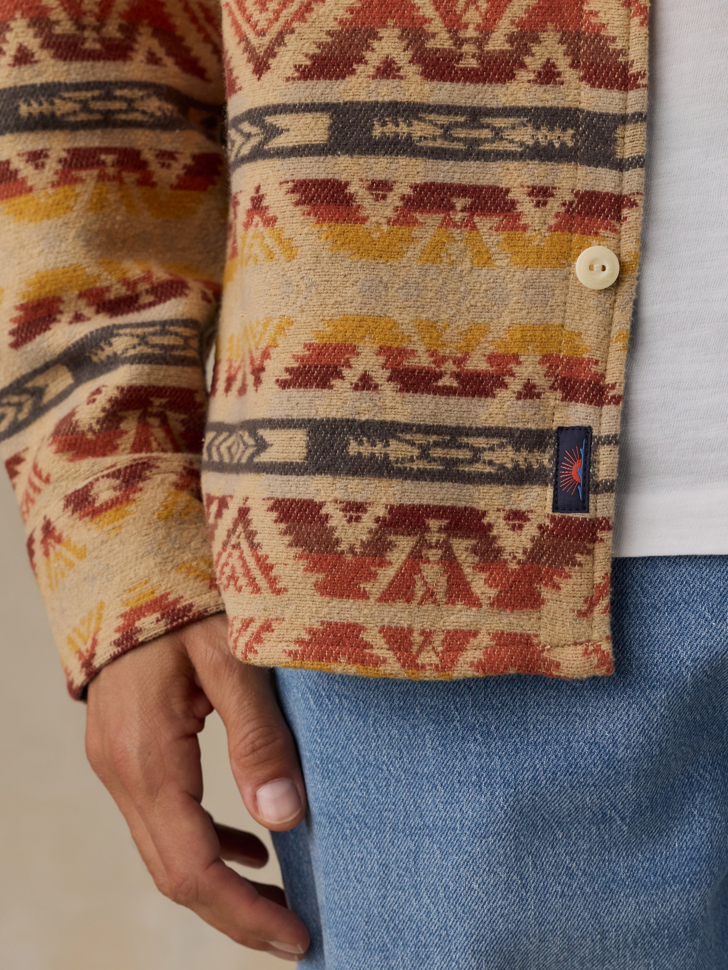 Close-up of a man wearing a patterned knit cardigan with a Southwestern geometric design in beige, rust red, mustard yellow, and dark gray. The cardigan has a cream-colored snap button and a small dark blue woven Faherty brand tag on the lower right cuff.