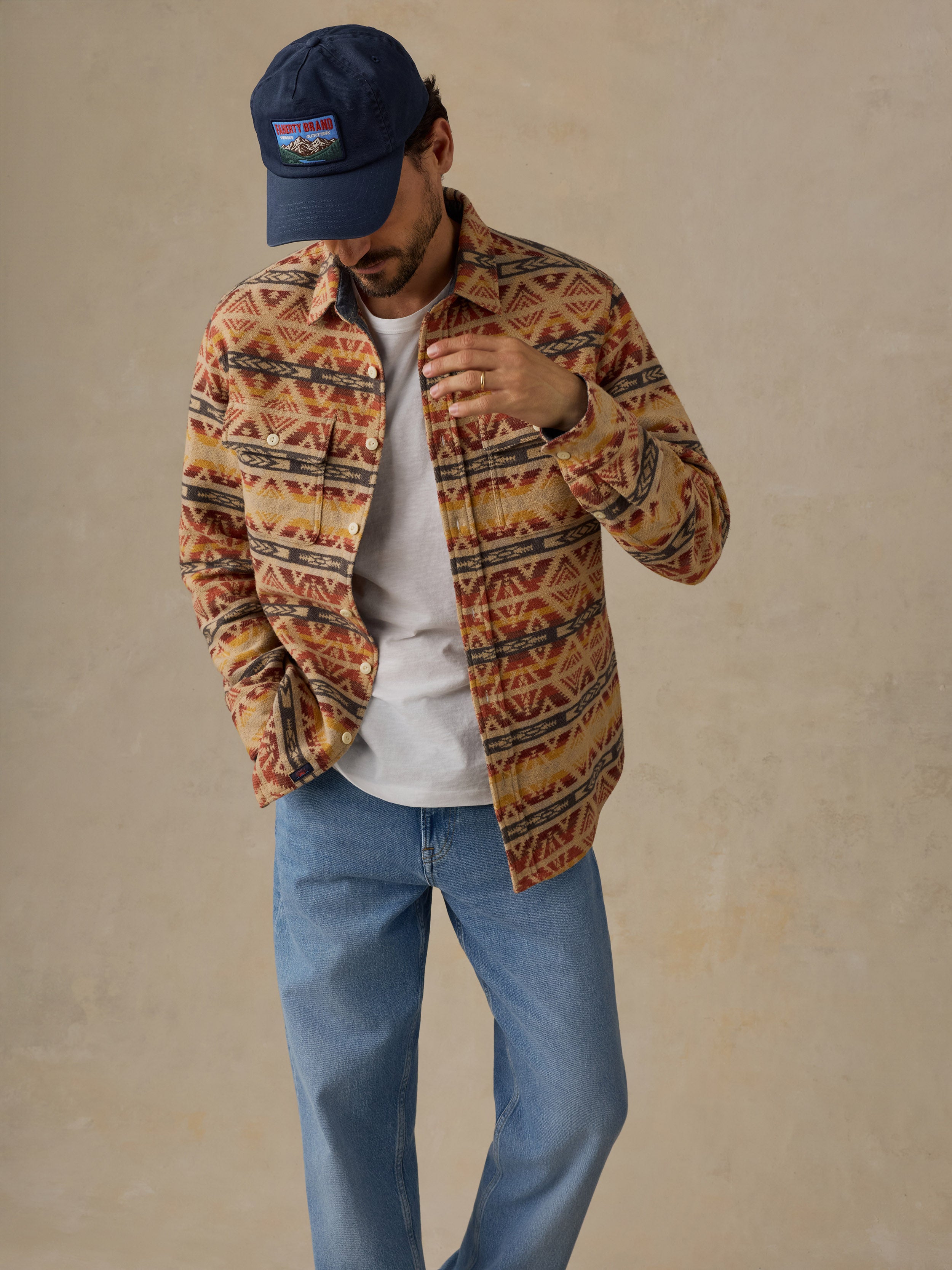 A man wearing a navy blue baseball cap, a brown and orange geometric patterned button-up shirt over a white t-shirt, and blue jeans, standing against a textured beige background.