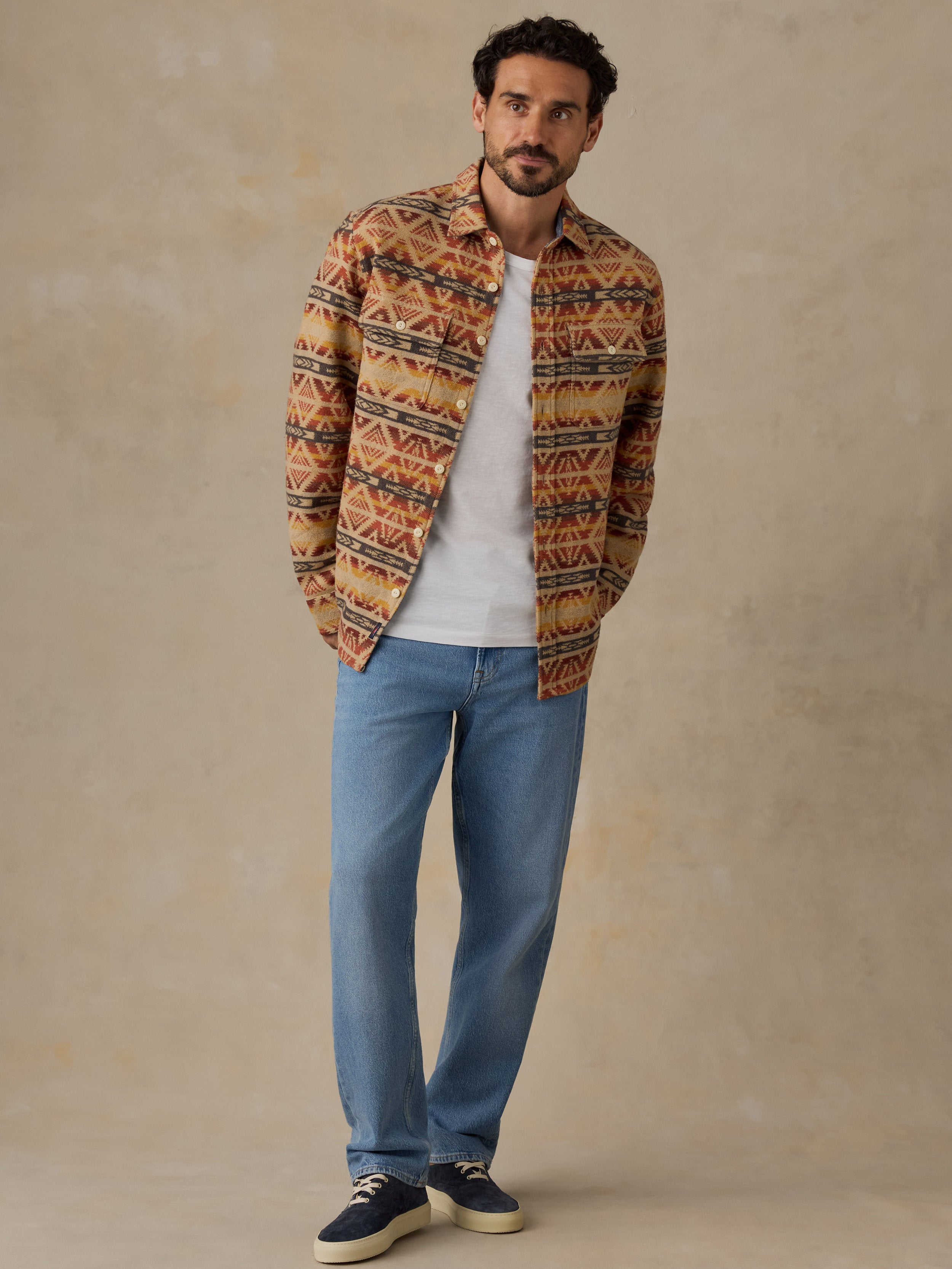 A man wearing a brown and orange geometric patterned button-up shirt over a white t-shirt, paired with blue jeans and navy sneakers, standing against a textured beige studio backdrop.
