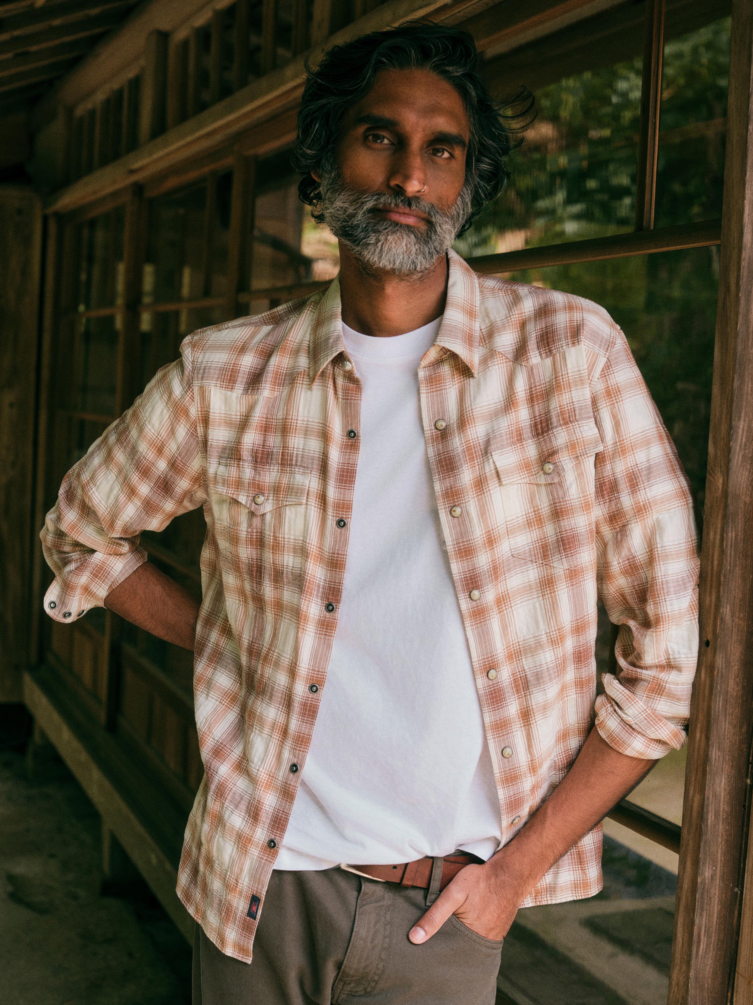 A man with a graying beard and dark hair stands on a rustic wooden porch wearing a light beige and brown plaid button-down shirt over a white t-shirt and khaki pants.