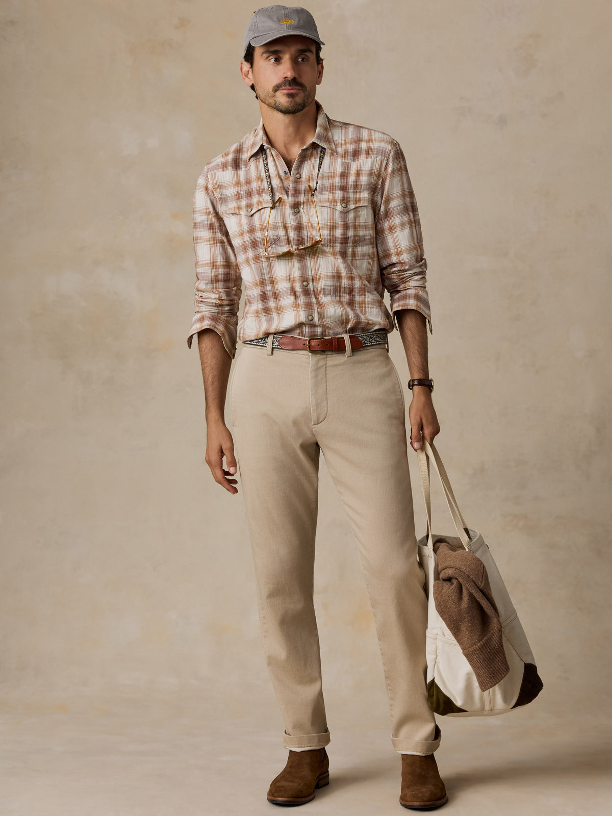 A man modeling a casual outdoor-style outfit featuring a brown and beige plaid button-down shirt, khaki pants, a gray baseball cap, and brown boots, while holding a canvas tote bag against a textured beige backdrop.