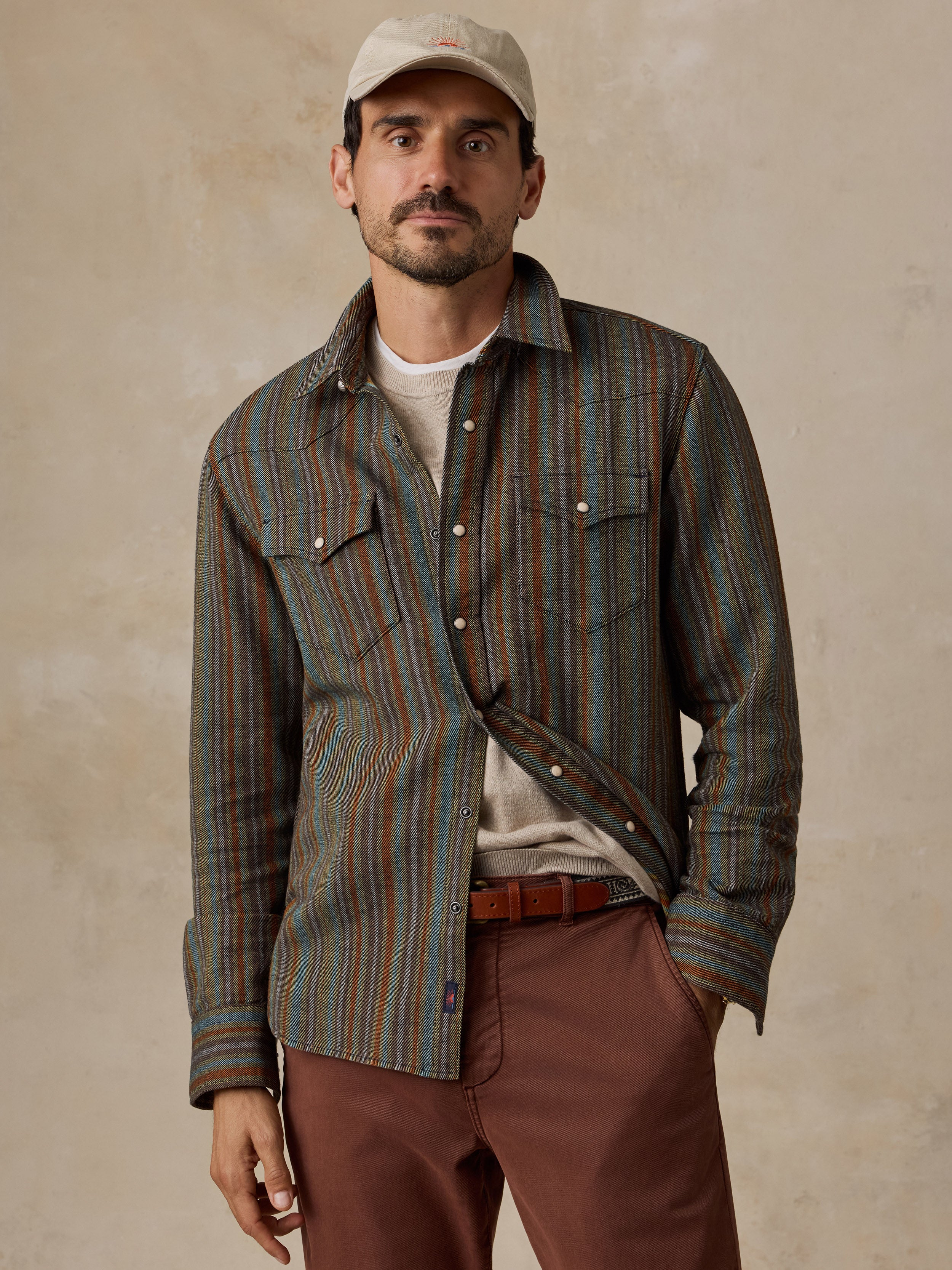 A man wearing a striped button-down shirt with blue, green, and brown vertical stripes, paired with brown pants and a beige baseball cap