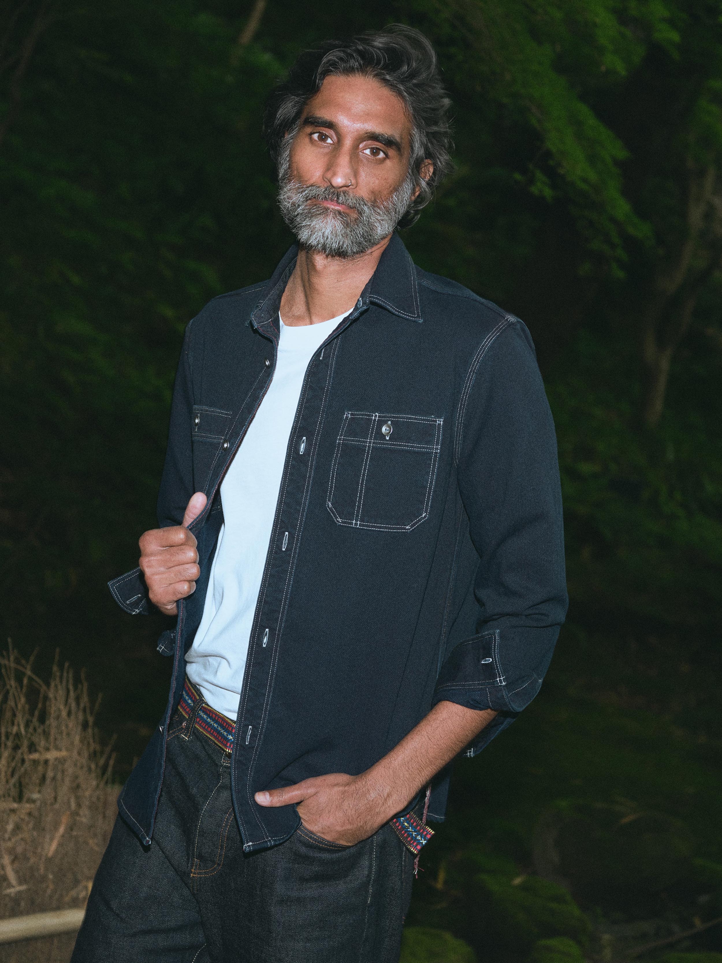 A man with a graying beard wearing a dark indigo denim button-up shirt over a white t-shirt and dark jeans, standing outdoors against a dark, blurry background of foliage.