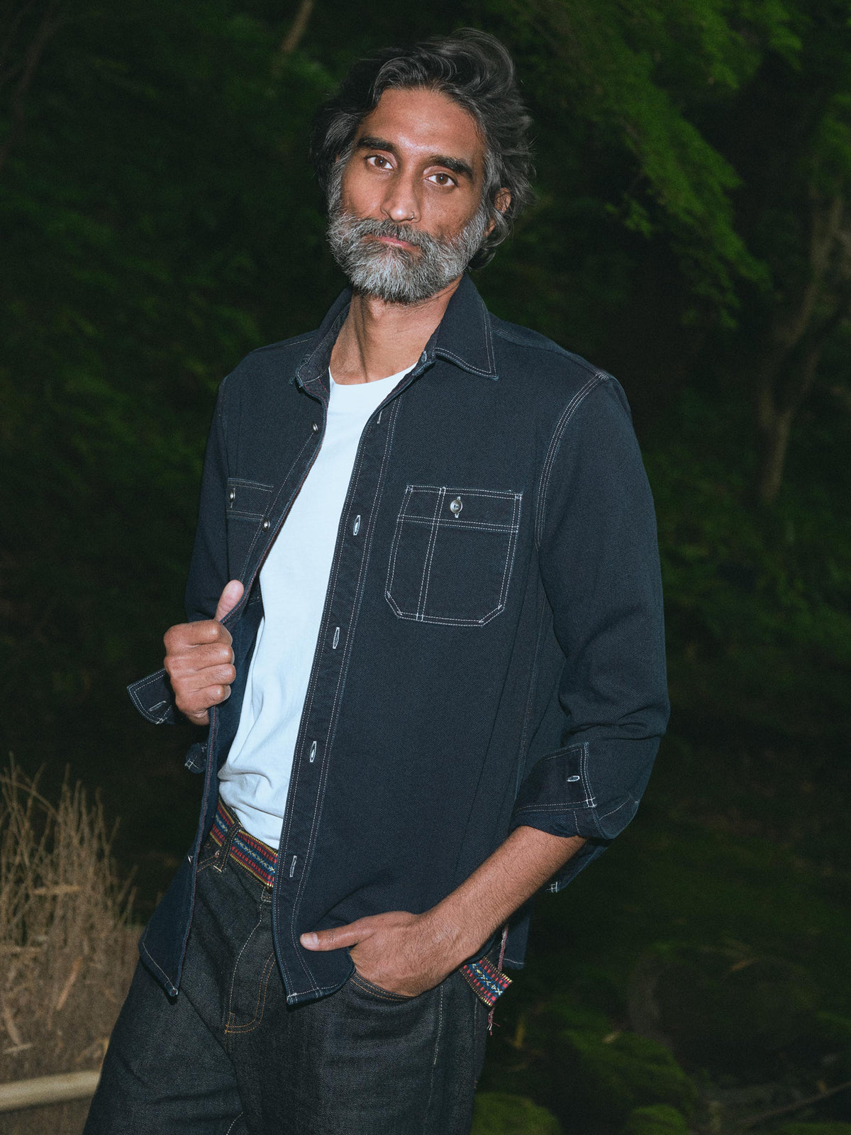 A man with a graying beard wearing a dark indigo denim button-up shirt over a white t-shirt and dark jeans, standing outdoors against a dark, blurry background of foliage., image 1 of 6