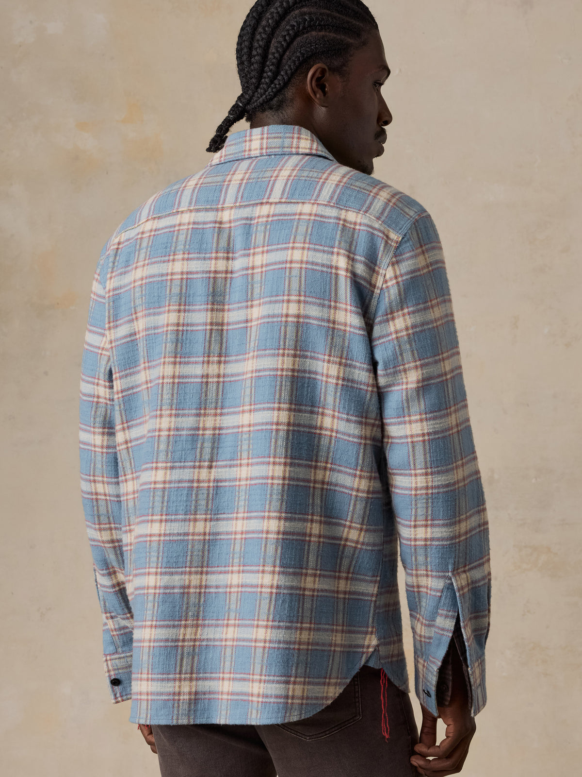 A man viewed from behind wearing a light blue plaid shirt over a plaid button up with brown pants, image 5 of 7