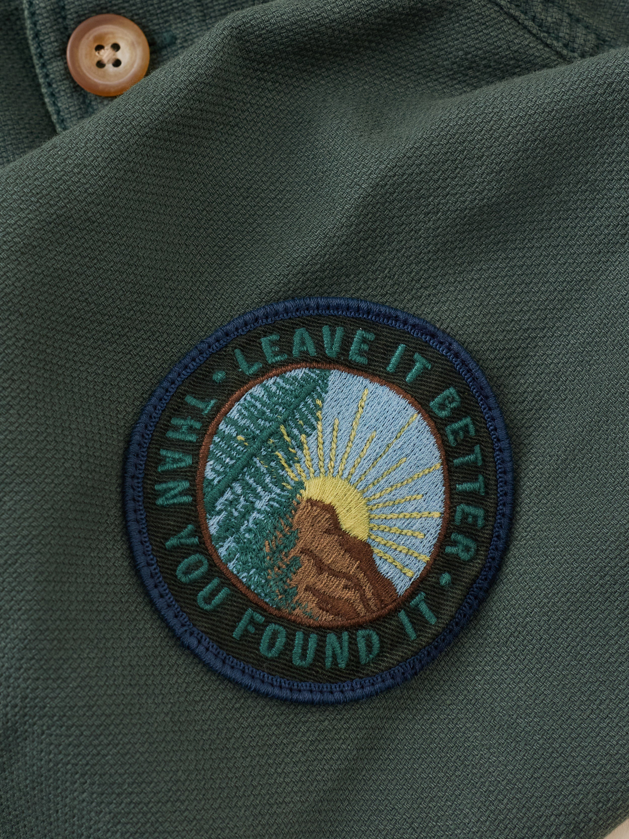 Close-up of an embroidered circular patch on dark olive green fabric, featuring a mountain and trees patch with a yellow sun and the text 'LEAVE IT BETTER THAN YOU LEFT IT'.