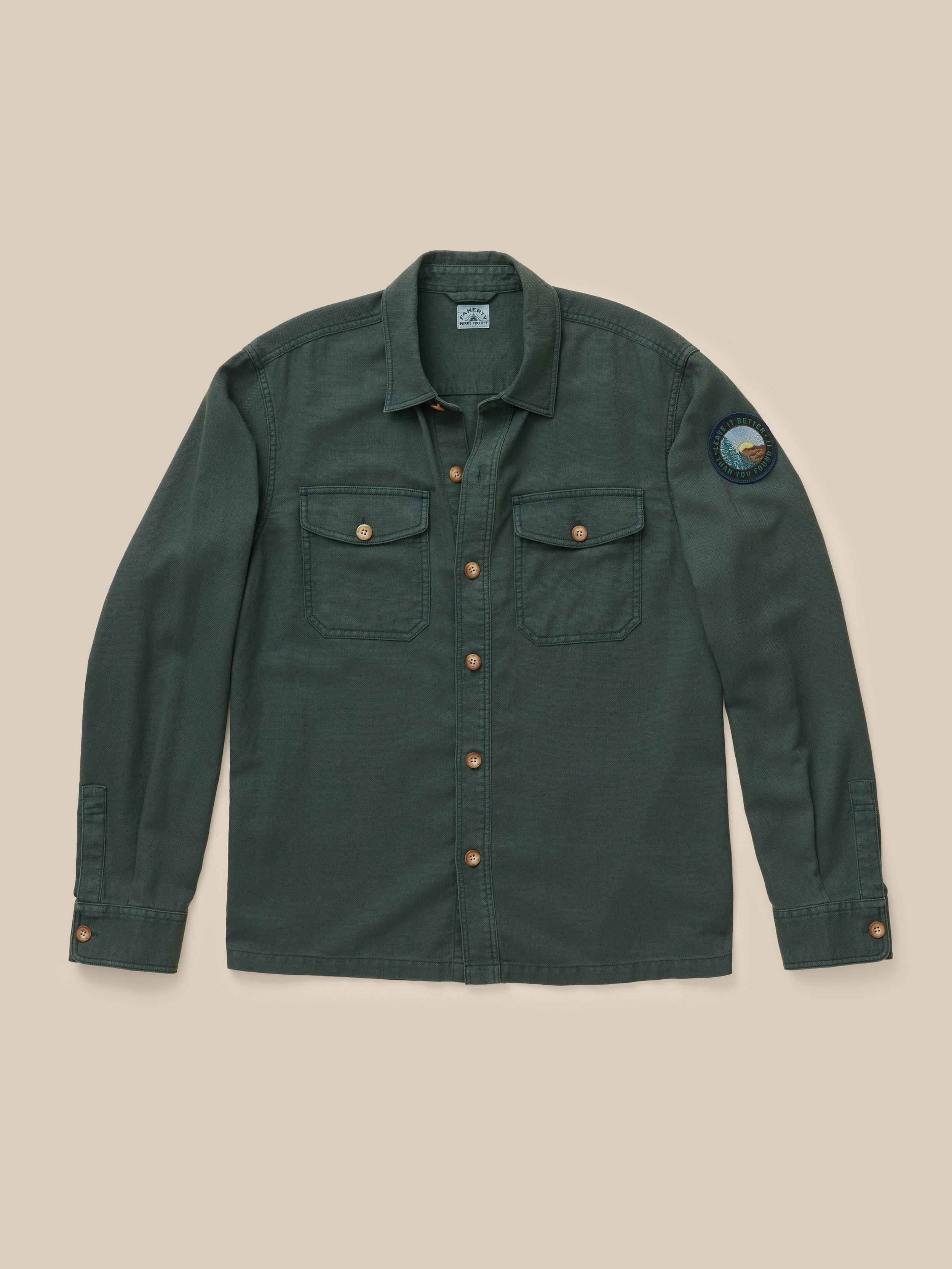 A dark green long-sleeve button-up shirt jacket with two chest flap pockets and a circular patch on the left sleeve, laid flat against a solid beige background.