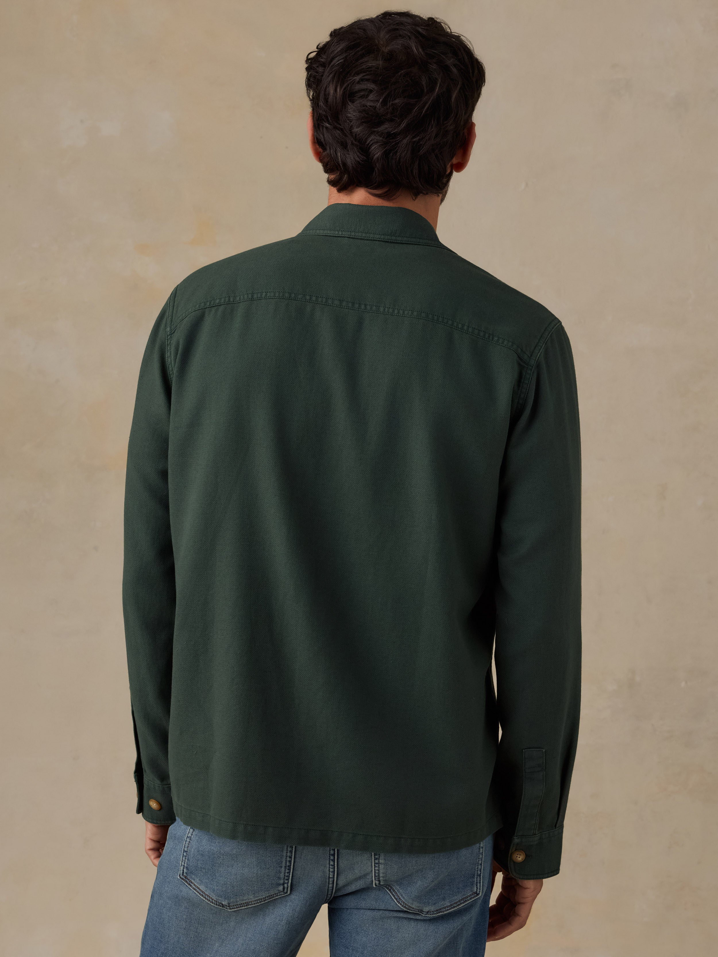 A man viewed from behind wearing a dark forest green long-sleeve button-up shirt and blue jeans, standing against a textured beige background.