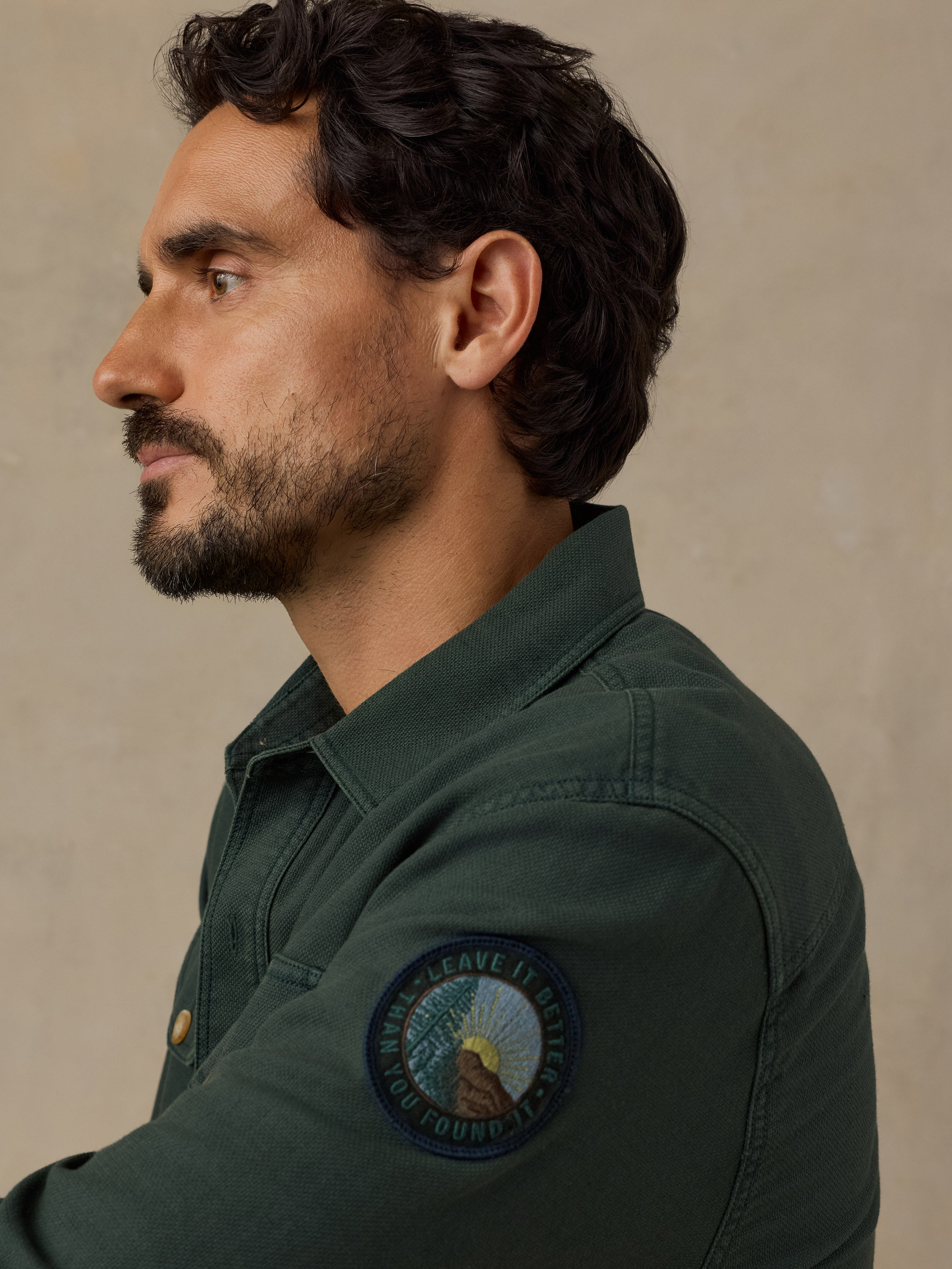 A man in profile wearing a dark green button-up shirt with a circular 'Leave It Better Than You Found It' patch on the left sleeve, set against a neutral beige background.