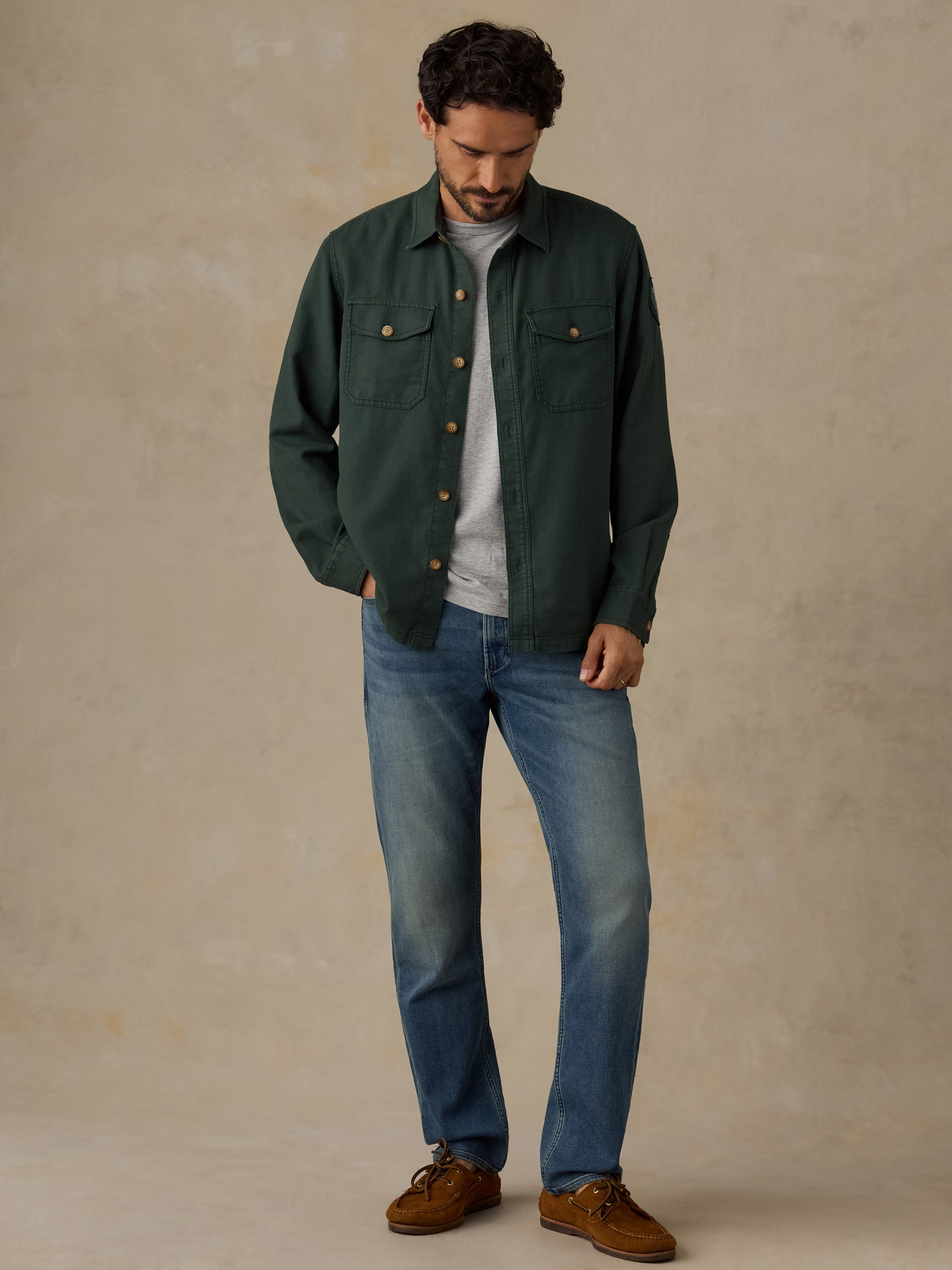 A man wearing a dark green button-up overshirt, blue jeans, and brown suede shoes, standing against a textured beige studio backdrop.