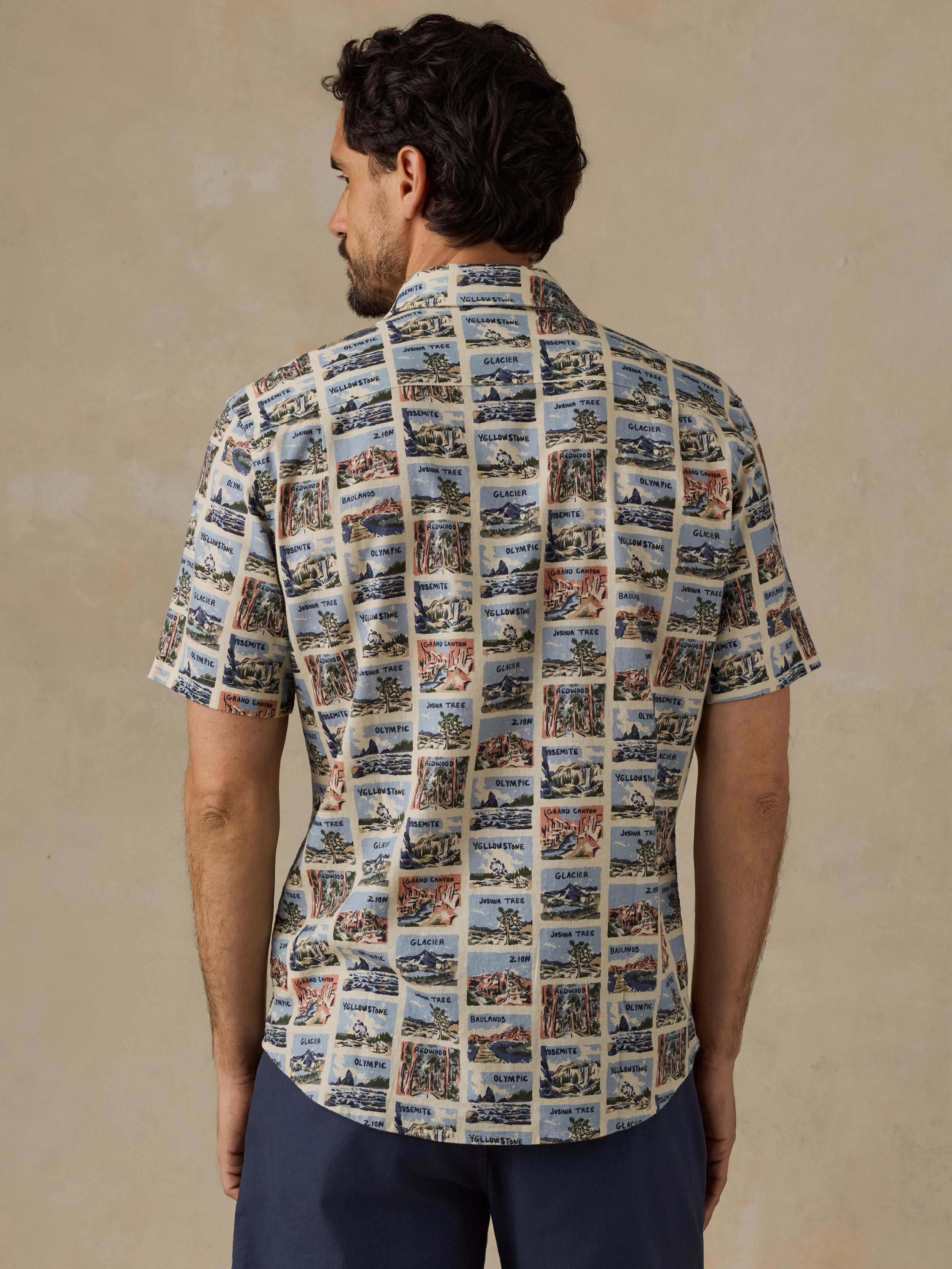 A man viewed from the back wearing a short-sleeve button-up shirt with an all-over collage print of vintage harbor scenes, paired with navy pants, against a textured beige studio background.