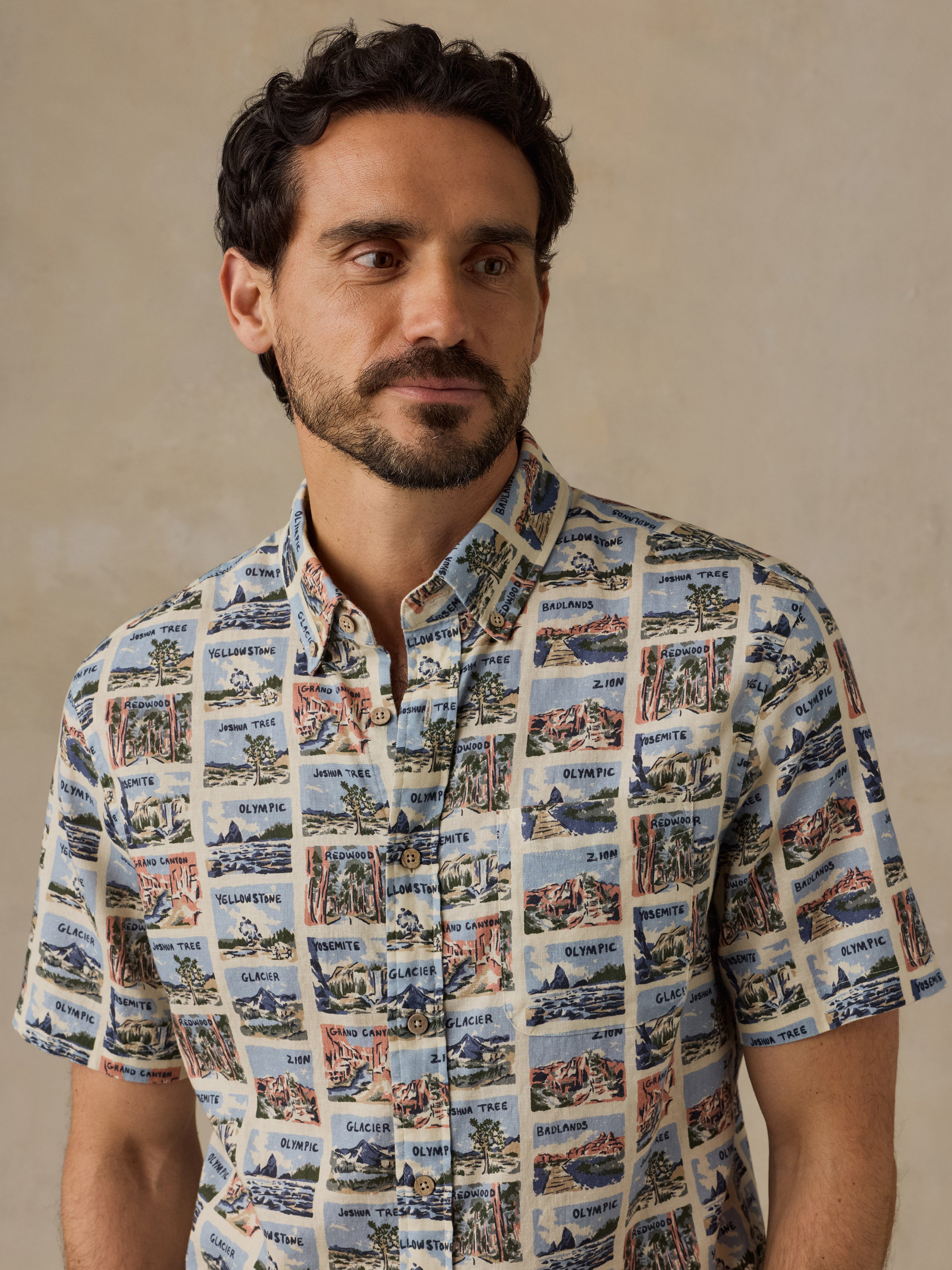 A man wearing a short-sleeve button-up shirt with a vintage postcard-style collage print featuring coastal scenes and text, posed against a neutral beige background.