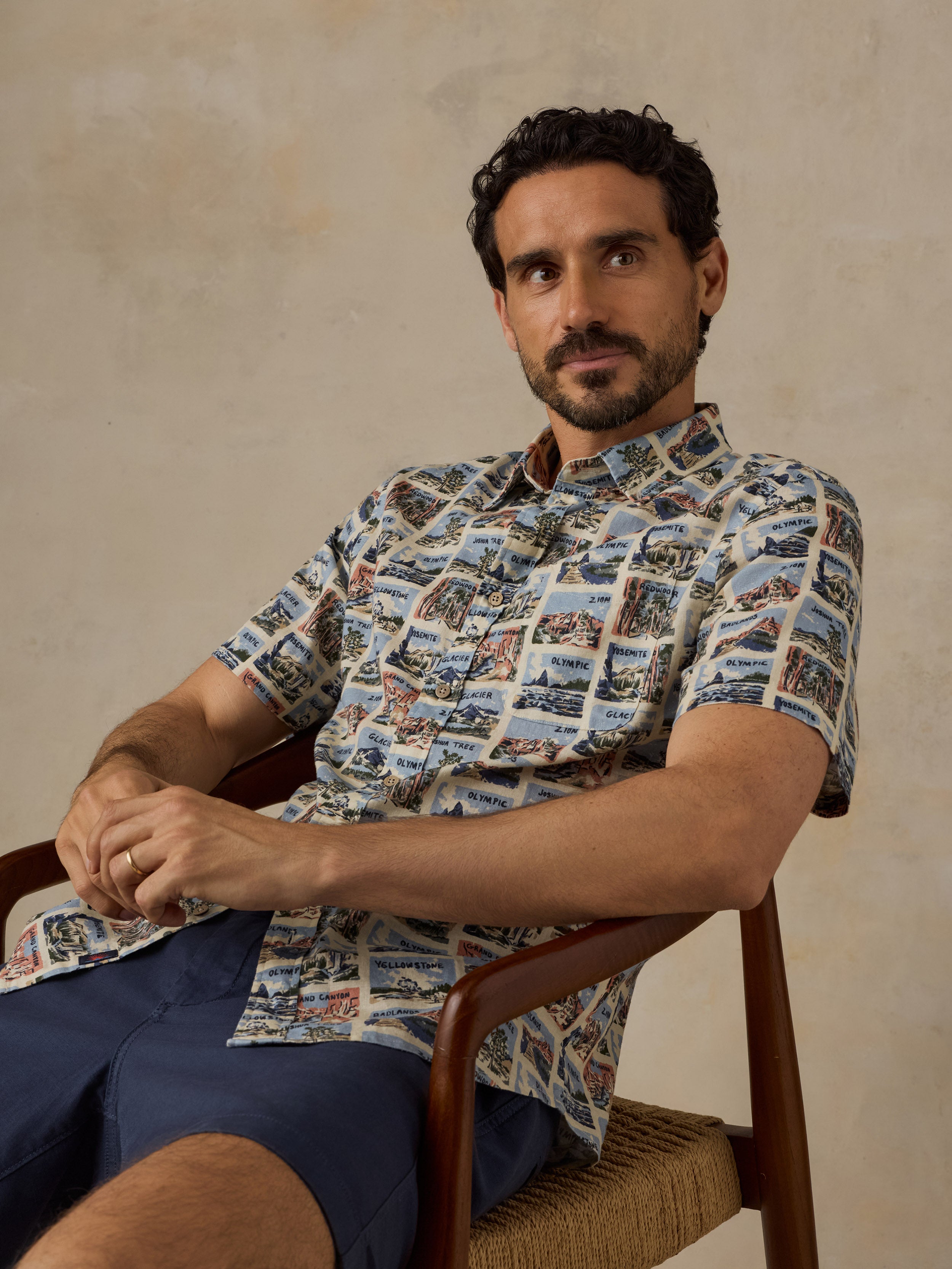 A man wearing a short-sleeve button-up shirt with a detailed scenic print, seated in a wooden chair against a textured beige wall.