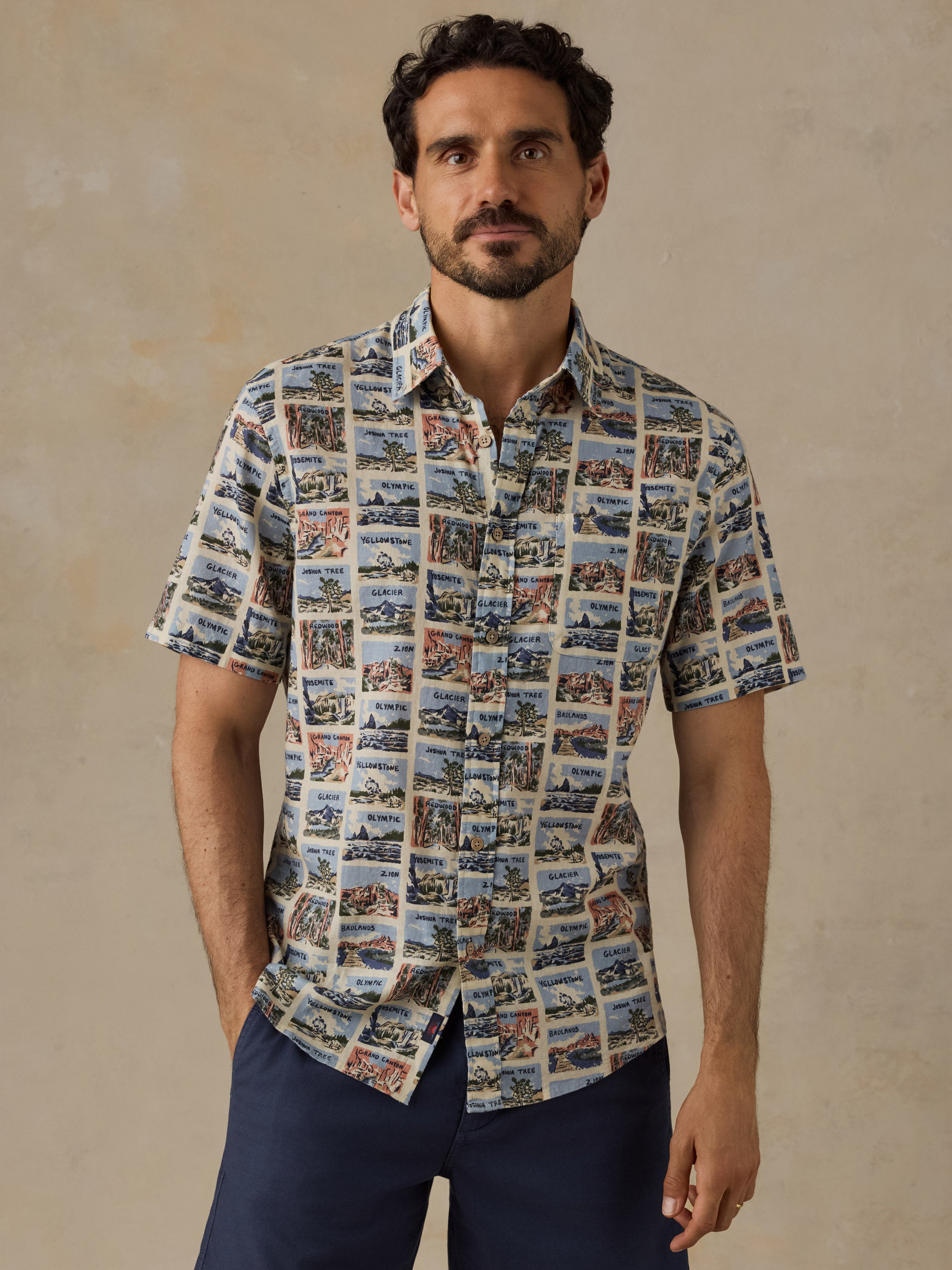 A man wearing a short-sleeve button-up shirt with a detailed scenic print, paired with navy blue pants, standing against a textured beige background.