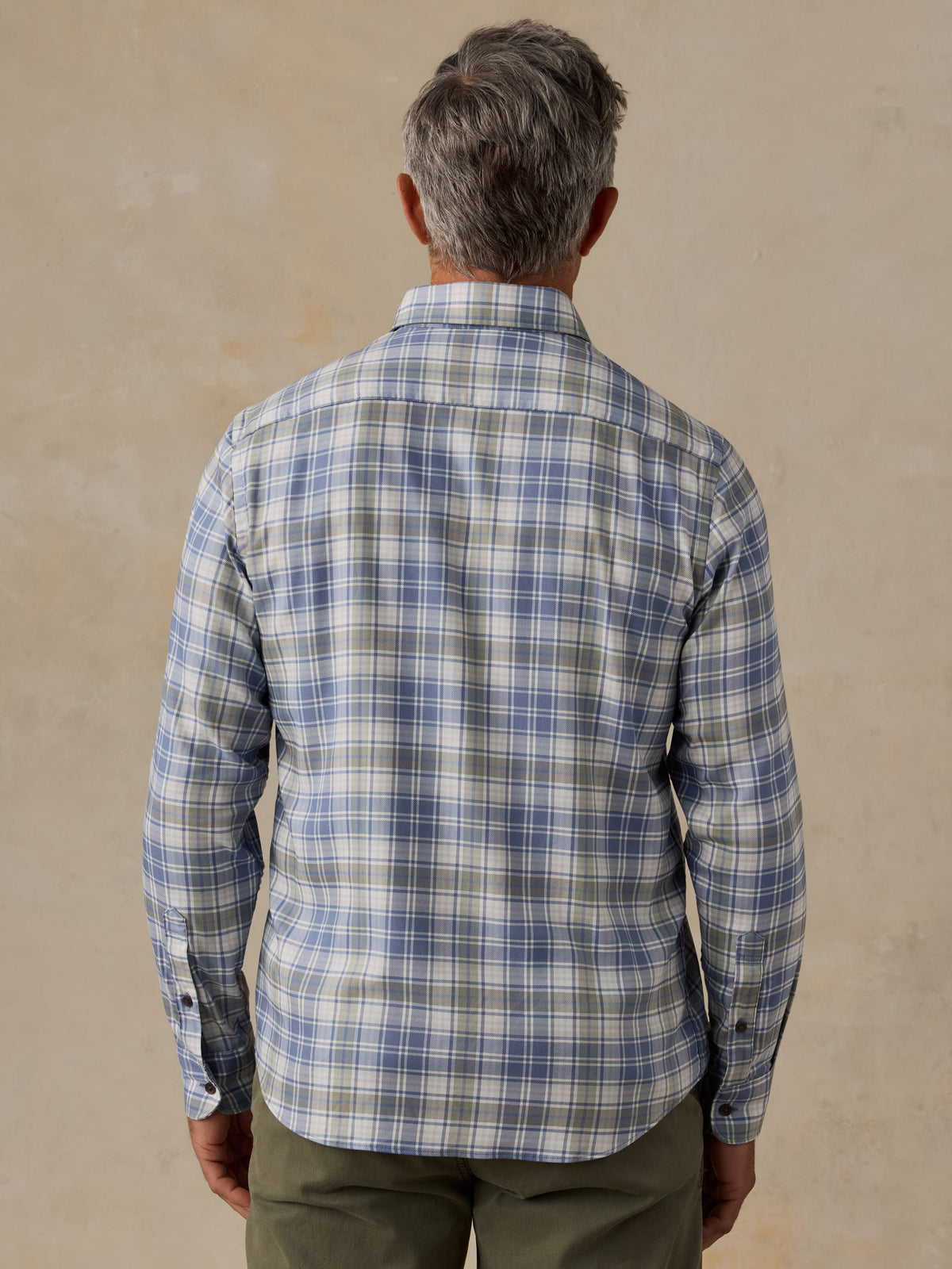 A man viewed from behind wearing a long-sleeve plaid button-down shirt in blue, gray, and white, paired with olive green pants, set against a textured beige wall., image 3 of 6