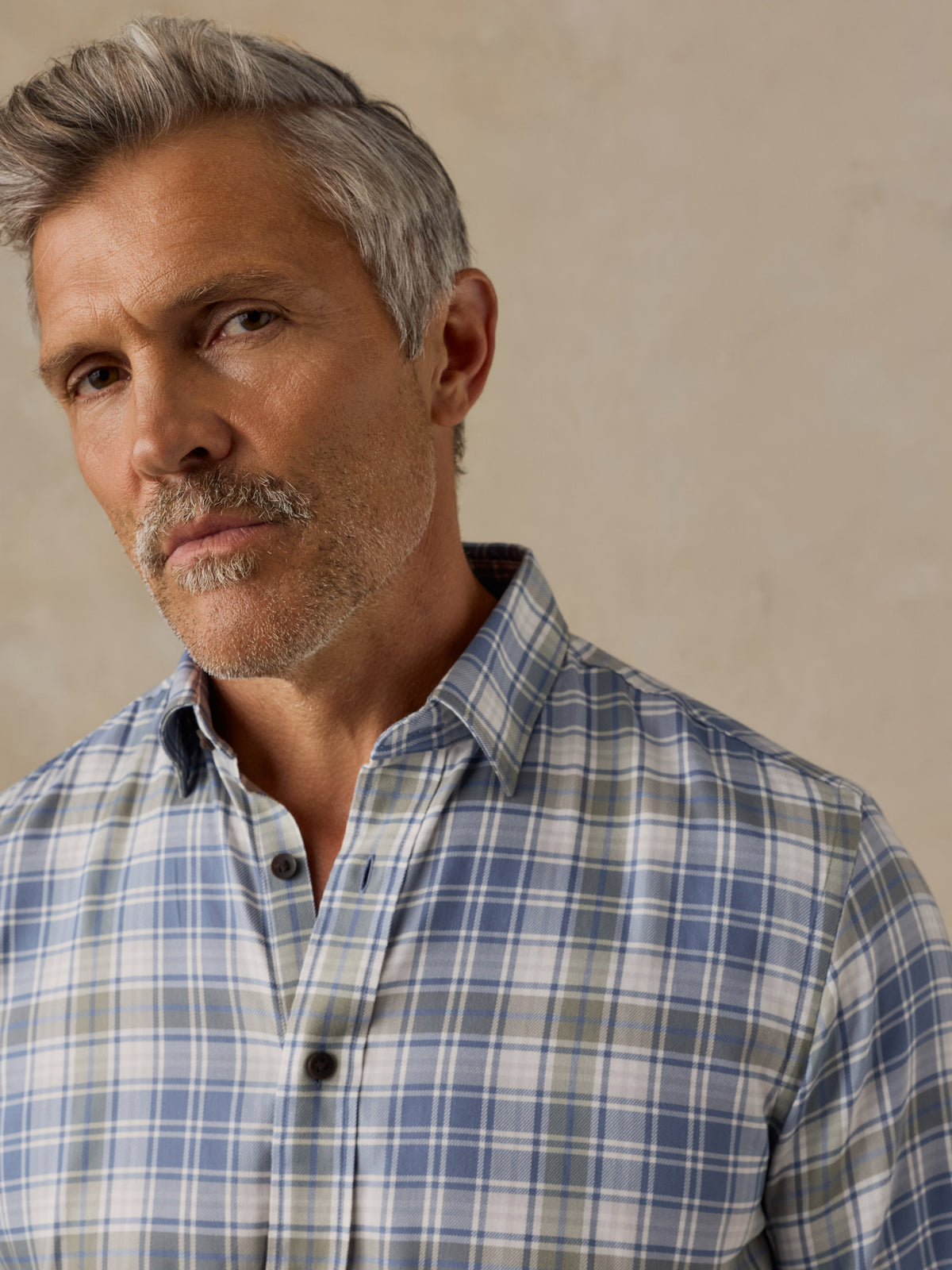 A close-up portrait of a man with gray hair and a mustache wearing a blue, white, and light green plaid button-down shirt against a textured beige background., image 2 of 6