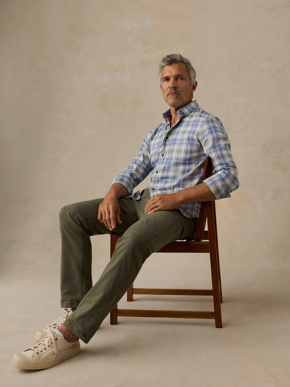 A man sits on a wooden chair wearing a light blue and white plaid button-down shirt, olive green pants, and cream-colored sneakers against a textured beige studio backdrop., image 5 of 6