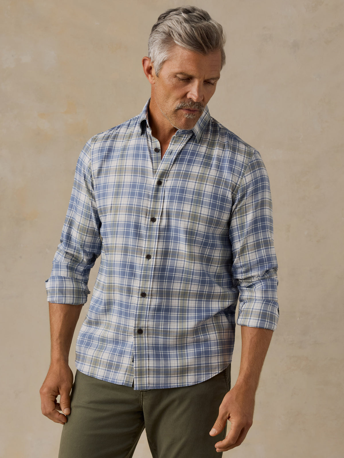 A man wearing a blue and white plaid button-down shirt with olive green pants, standing against a textured beige background., image 1 of 6