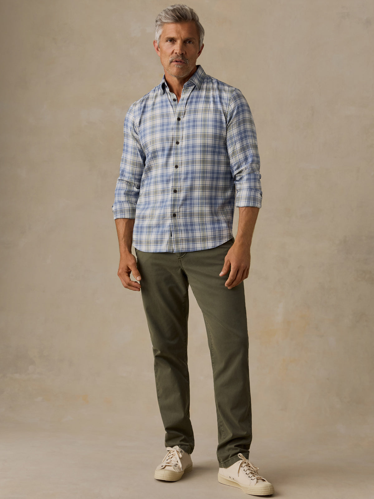 A man wearing a light blue and white plaid button-down shirt, olive green trousers, and off-white sneakers, standing against a textured beige studio backdrop., image 6 of 6