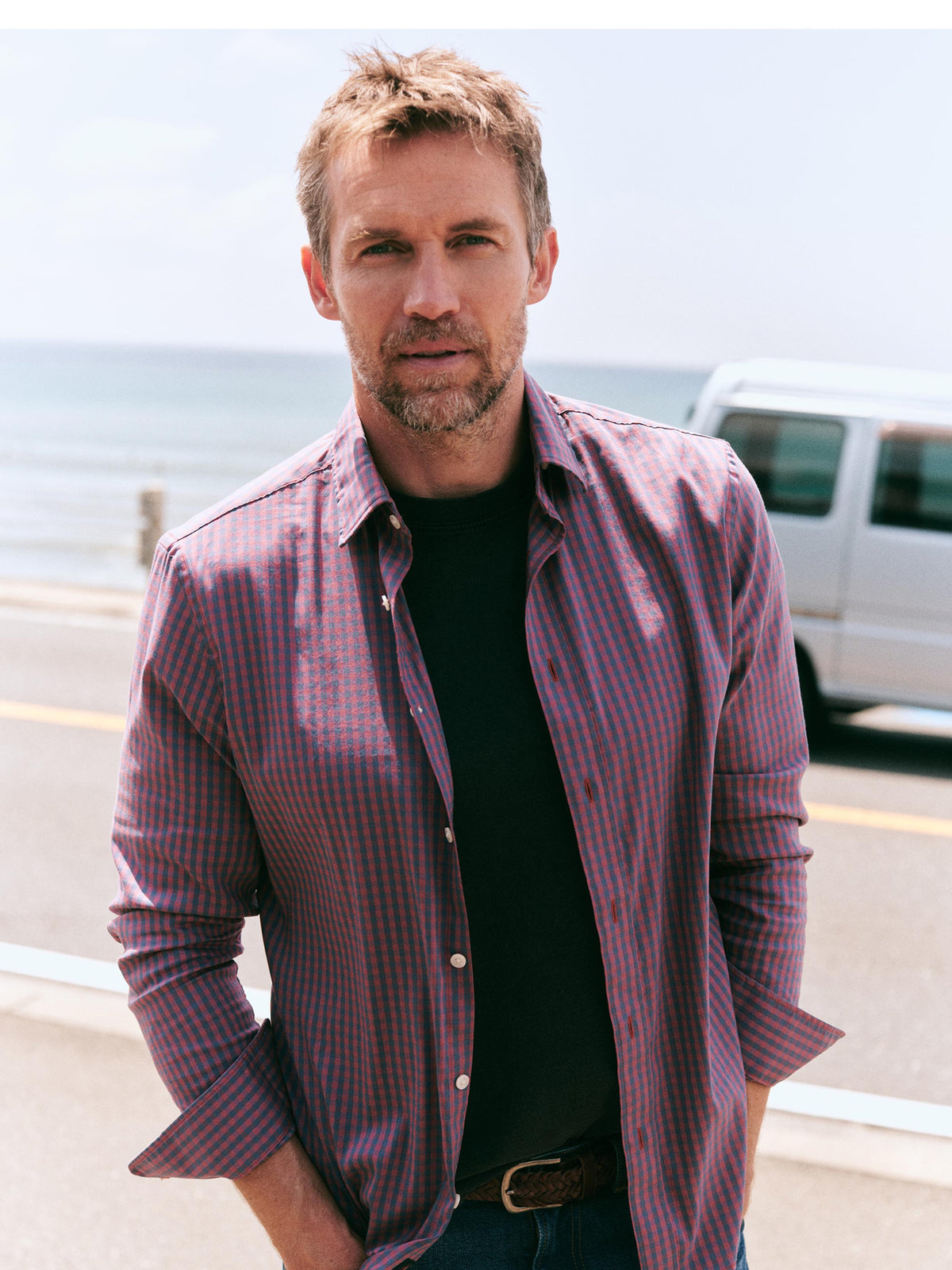 A man standing outdoors near the ocean wearing a purple and red striped button-down shirt over a black t-shirt, with a white van in the background.