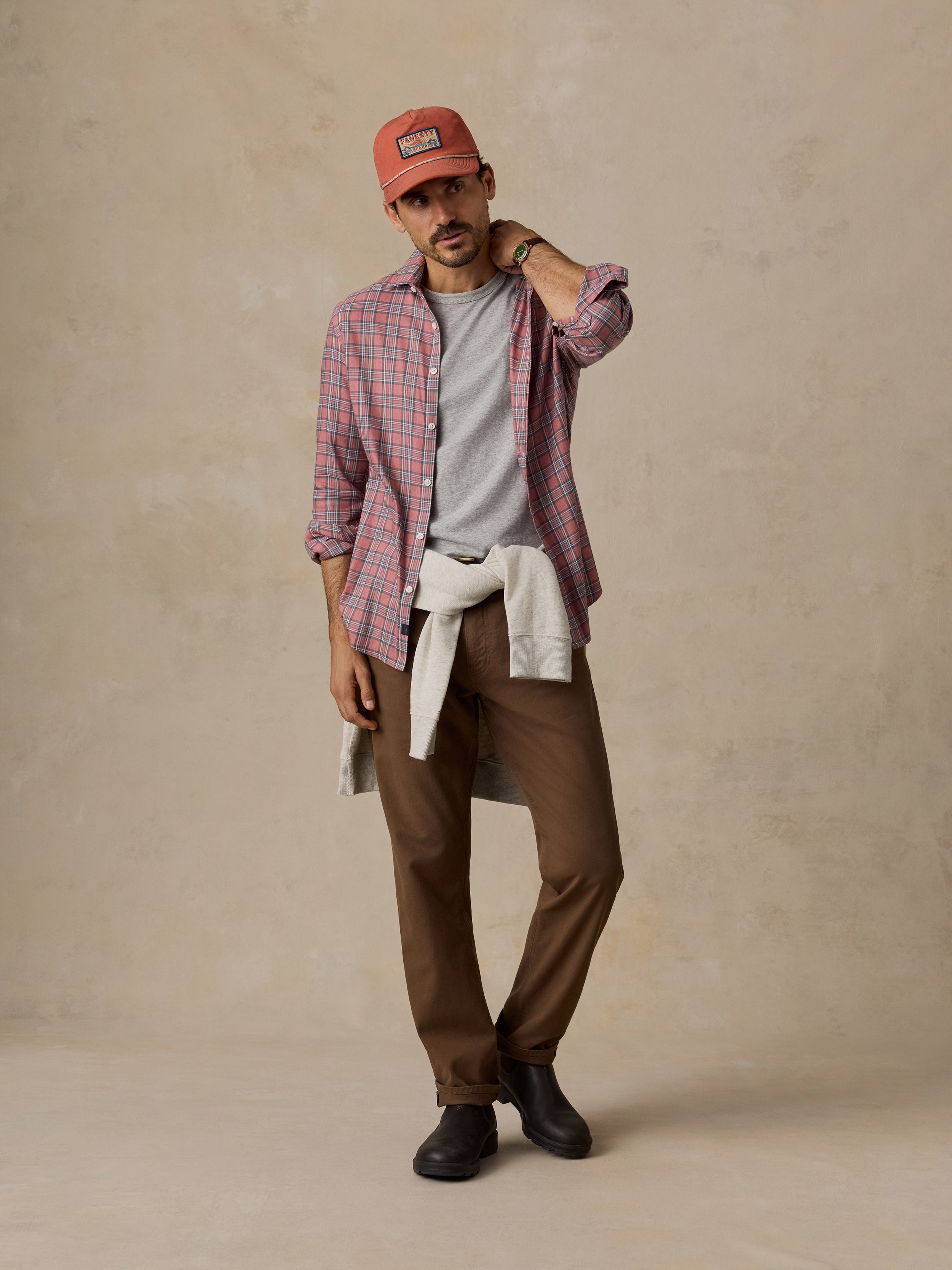 A man wearing a red and blue plaid button-down shirt, a gray t-shirt, brown trousers, and an orange baseball cap.