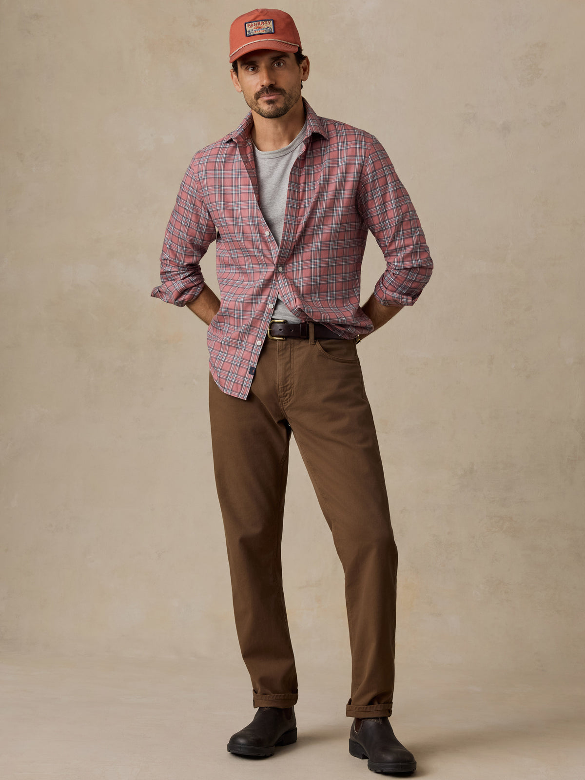 A man wearing a casual outfit consisting of a red and blue plaid button-down shirt, a light gray t-shirt, brown trousers, and an orange cap.