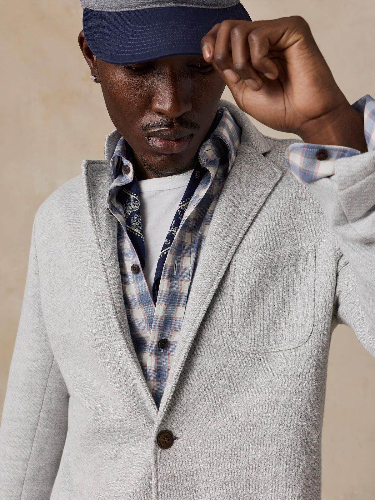 A man wearing a light gray wool-blend blazer over a blue and white plaid button-down shirt, over a white t-shirt and a navy baseball cap.