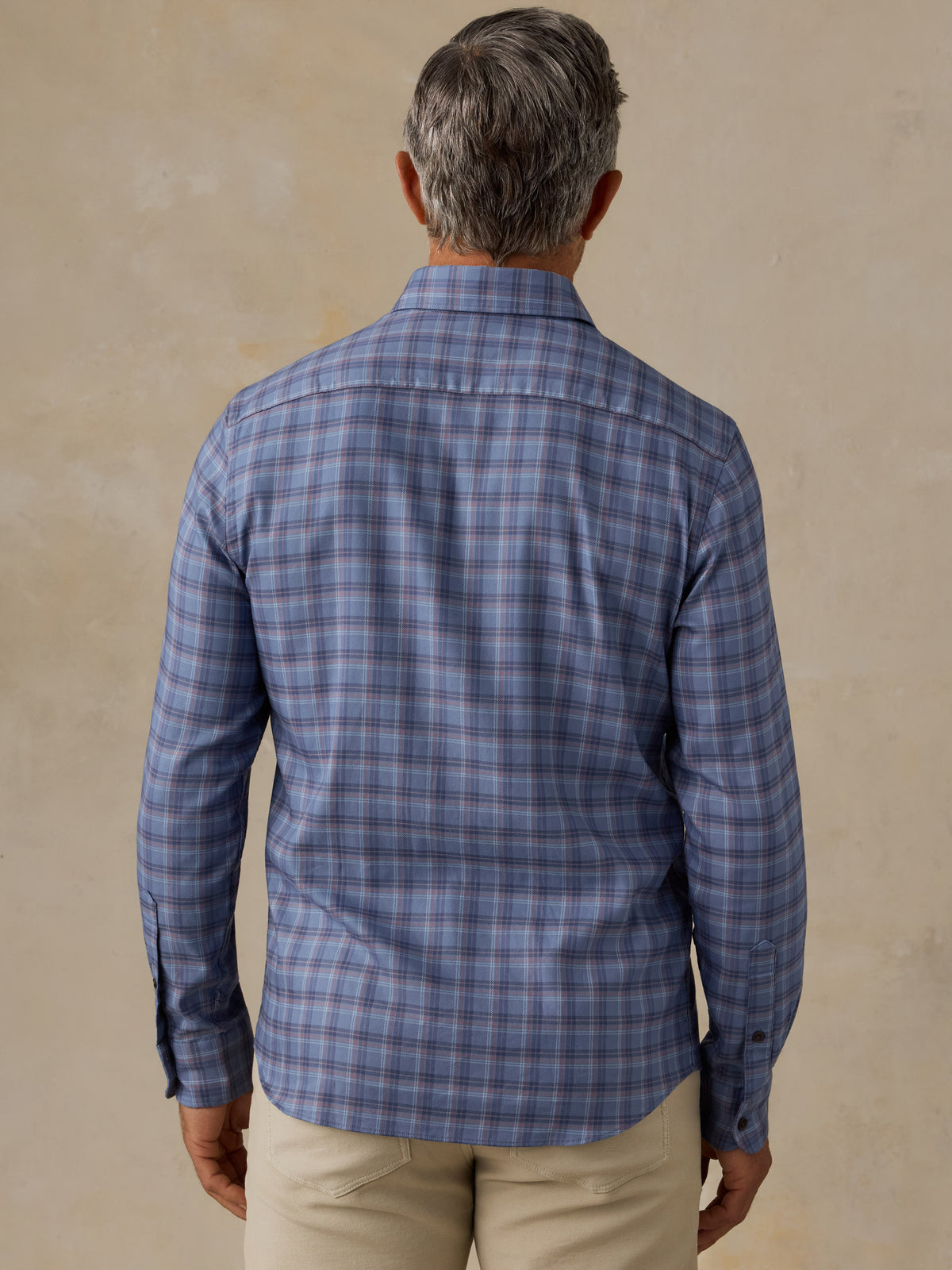 A man viewed from behind wearing a blue gray plaid long-sleeve button-down shirt and khaki pants, standing against a textured beige wall., image 3 of 6