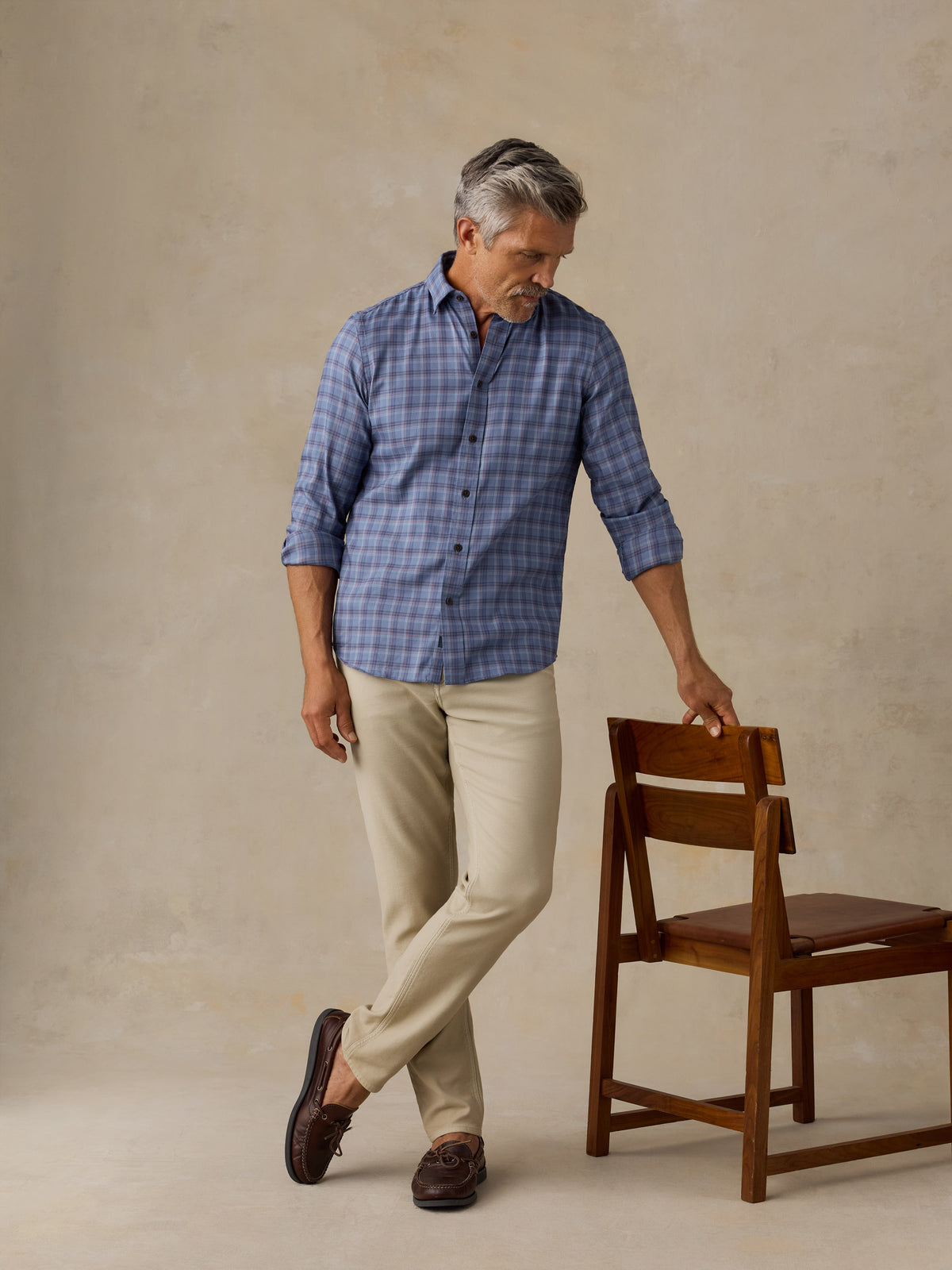 A man wearing a blue gray plaid button-down shirt, beige trousers, and brown loafers stands next to a wooden chair in a studio with a textured beige background., image 4 of 6