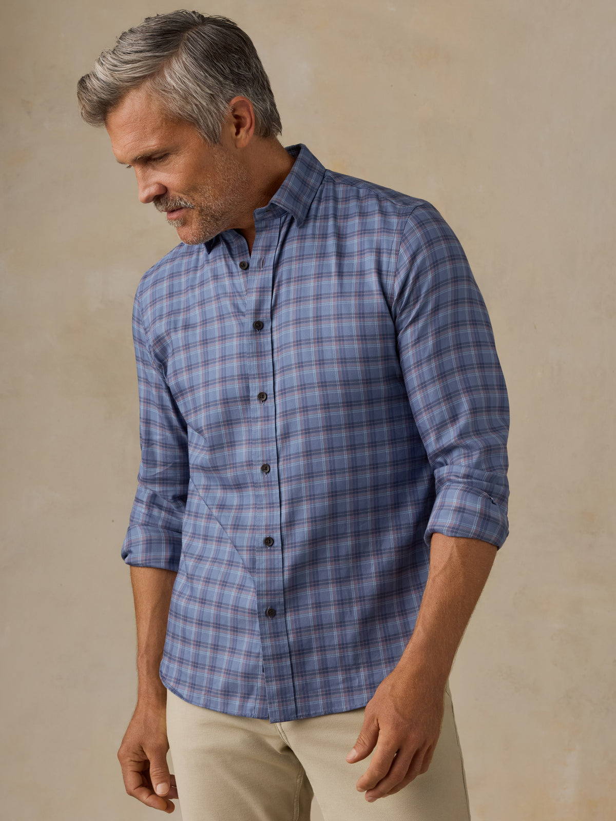 A man wearing a blue gray plaid button-down shirt with beige pants, standing against a textured beige background., image 1 of 6