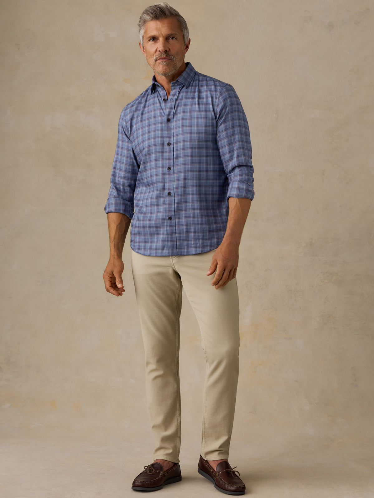 A man modeling a blue grey plaid button-down shirt with beige trousers and brown leather shoes, standing against a textured beige studio backdrop., image 6 of 6