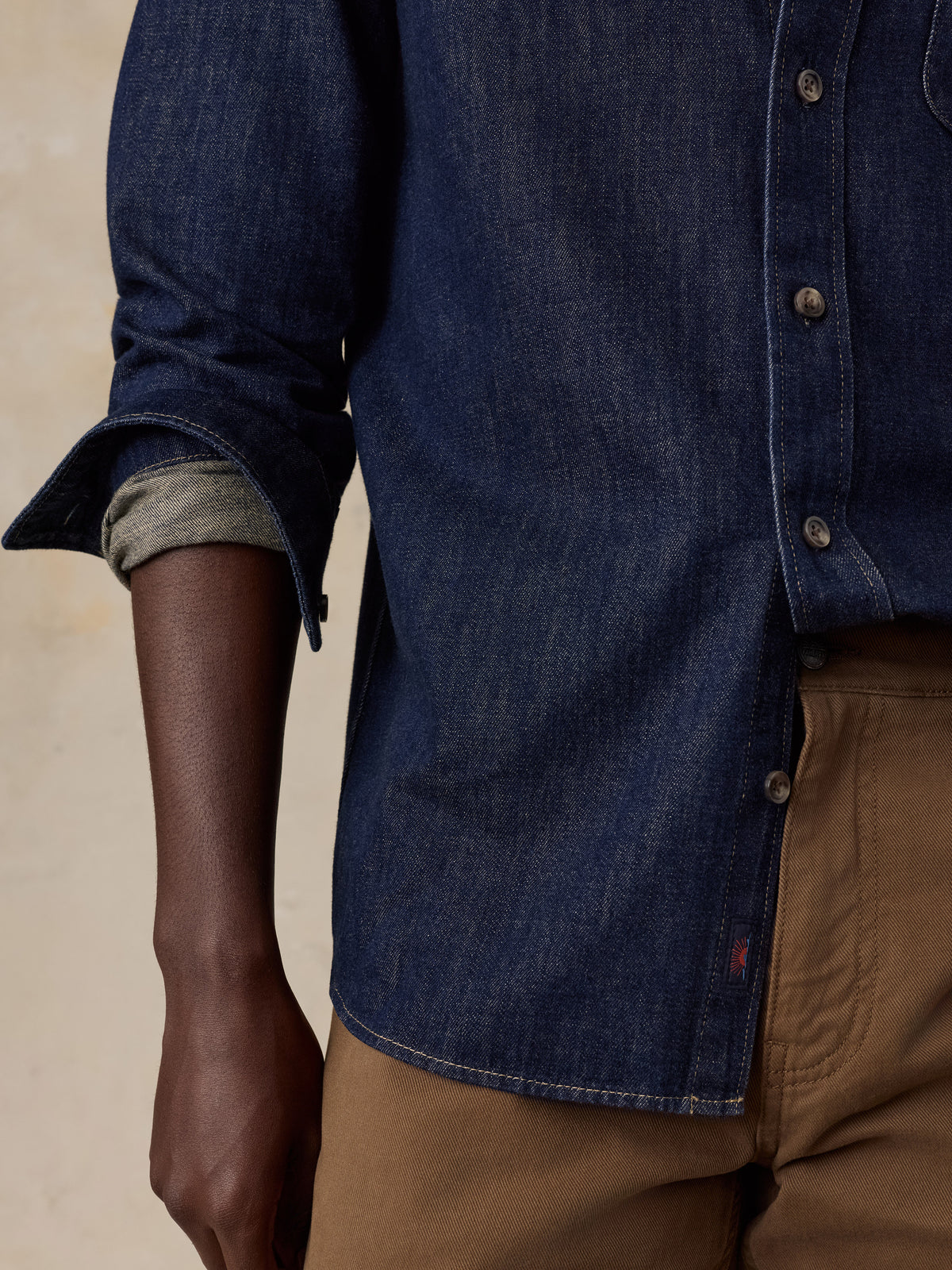 Close-up of a man wearing a dark indigo denim button-up shirt with rolled sleeves and brown pants, against a warm beige background.