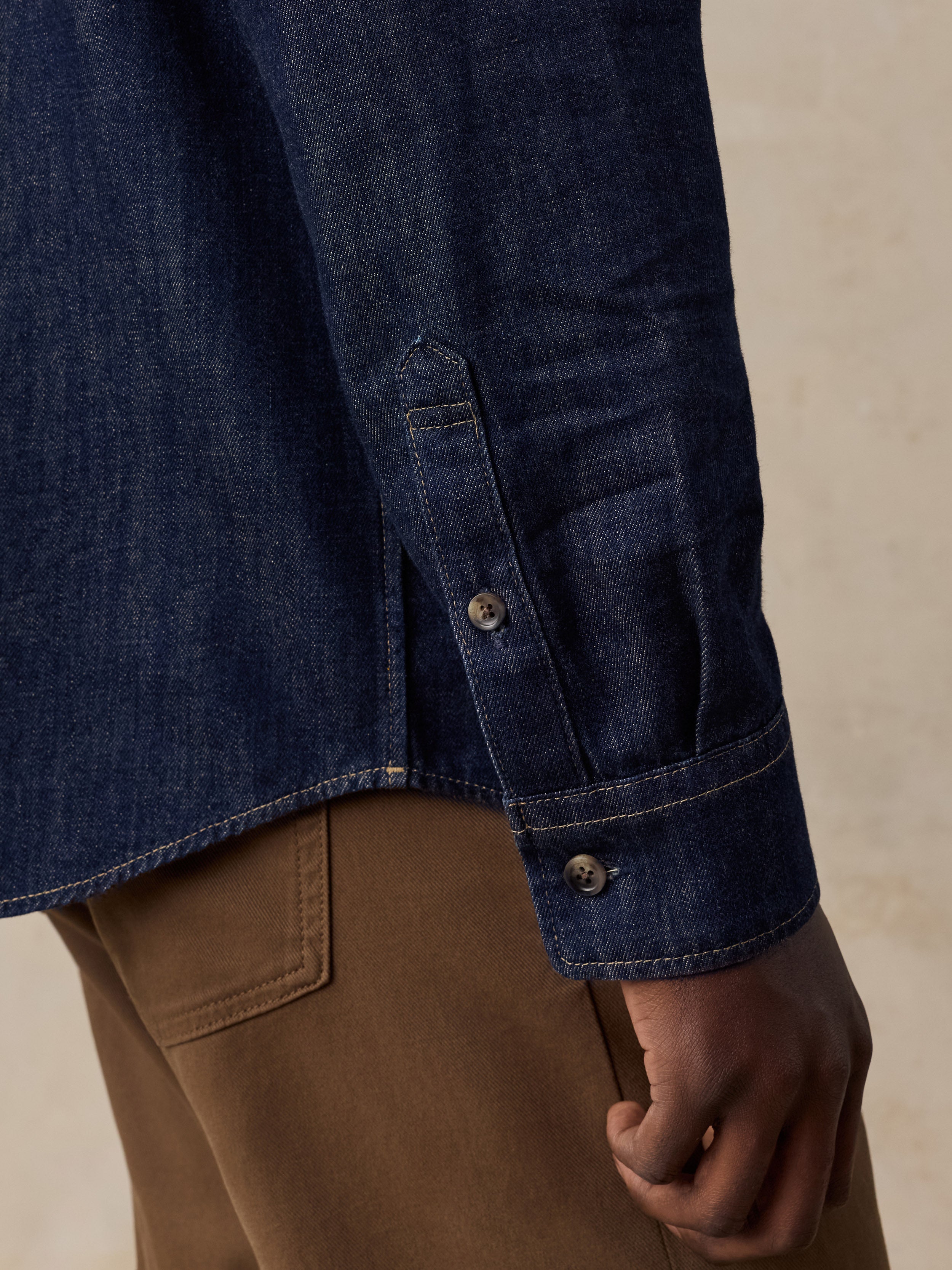 Close-up of a dark indigo denim shirt with a buttoned cuff pocket, worn by a model with brown trousers against a neutral beige background.