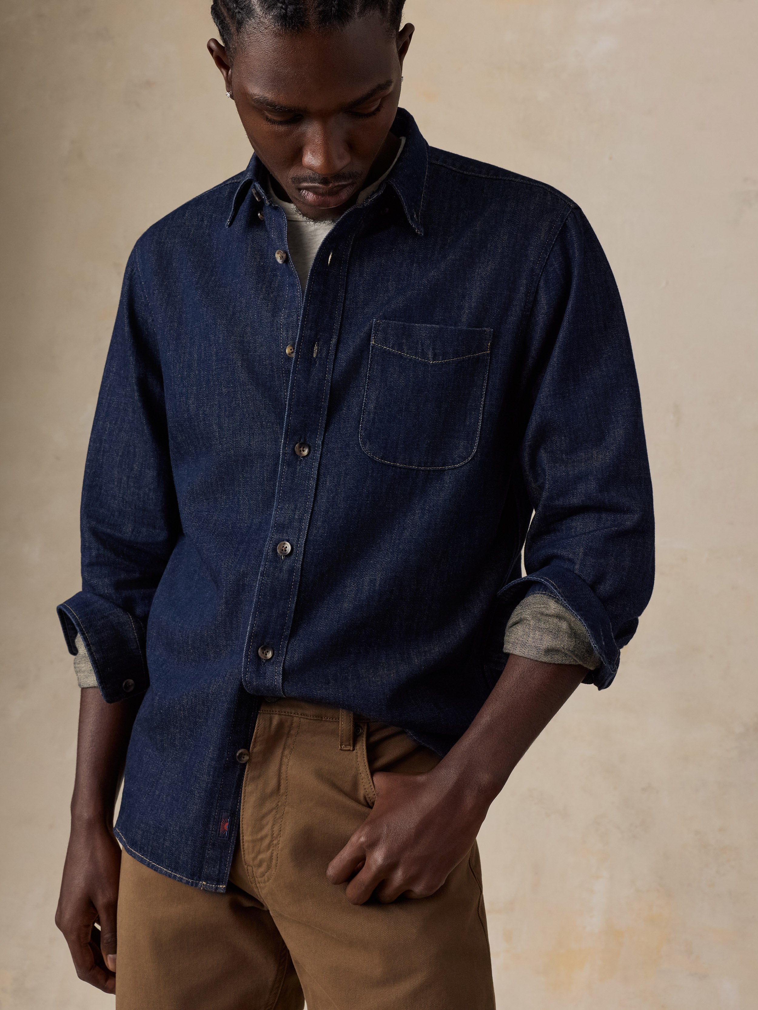 A man wearing a dark indigo denim long-sleeve button-up shirt with rolled-up sleeves, paired with brown trousers, standing against a textured beige background.