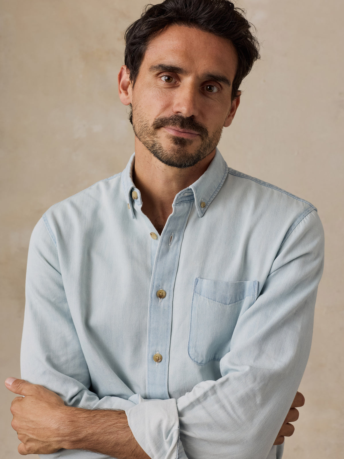 A man wearing a light blue long-sleeve button-down shirt with buttons, posed against a neutral beige textured background., image 2 of 6