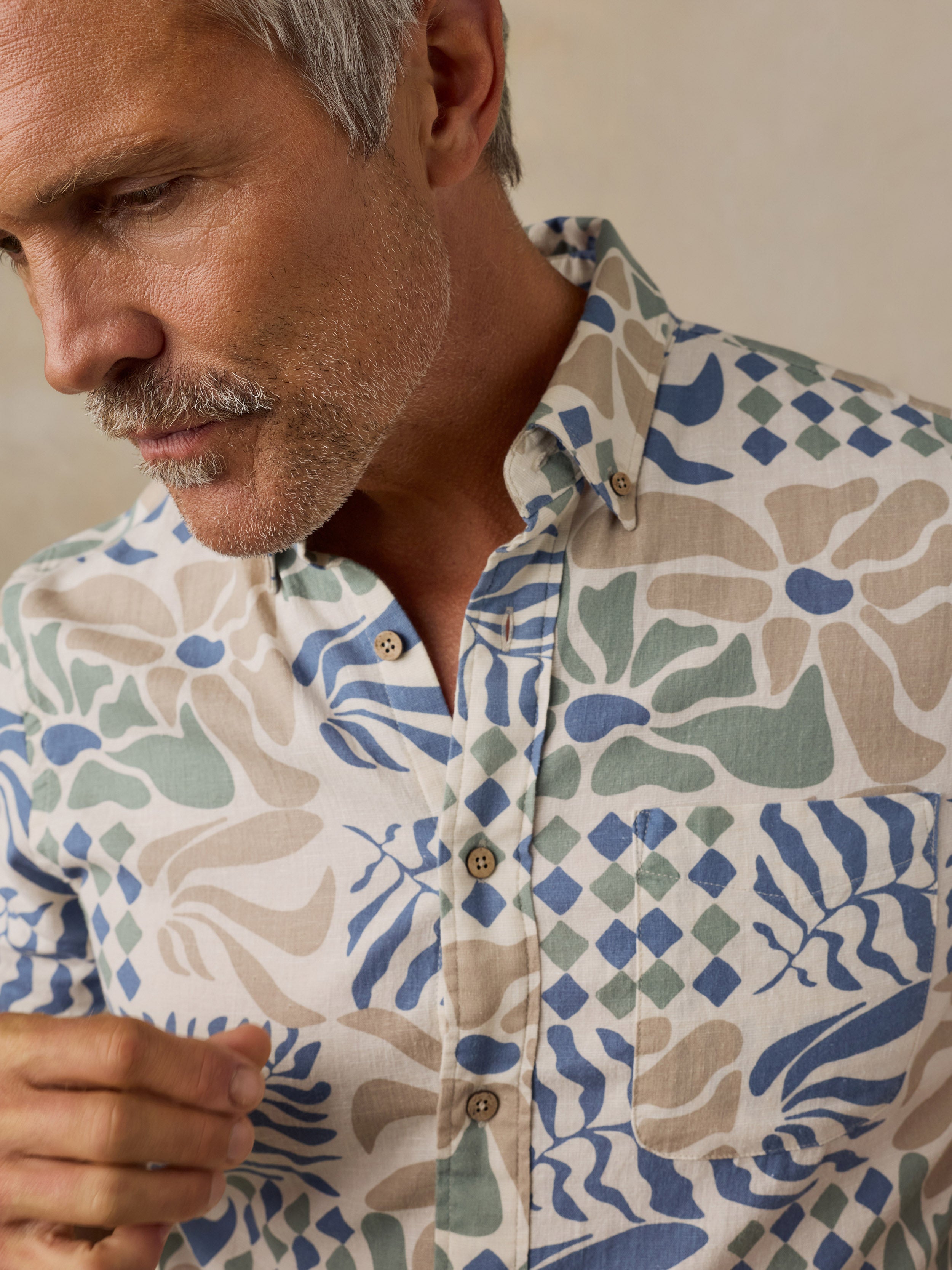 A close-up of a man wearing a short-sleeve button-up shirt with a beige, blue, and green botanical pattern. The man has gray hair and a mustache, and the background is a soft, warm beige.