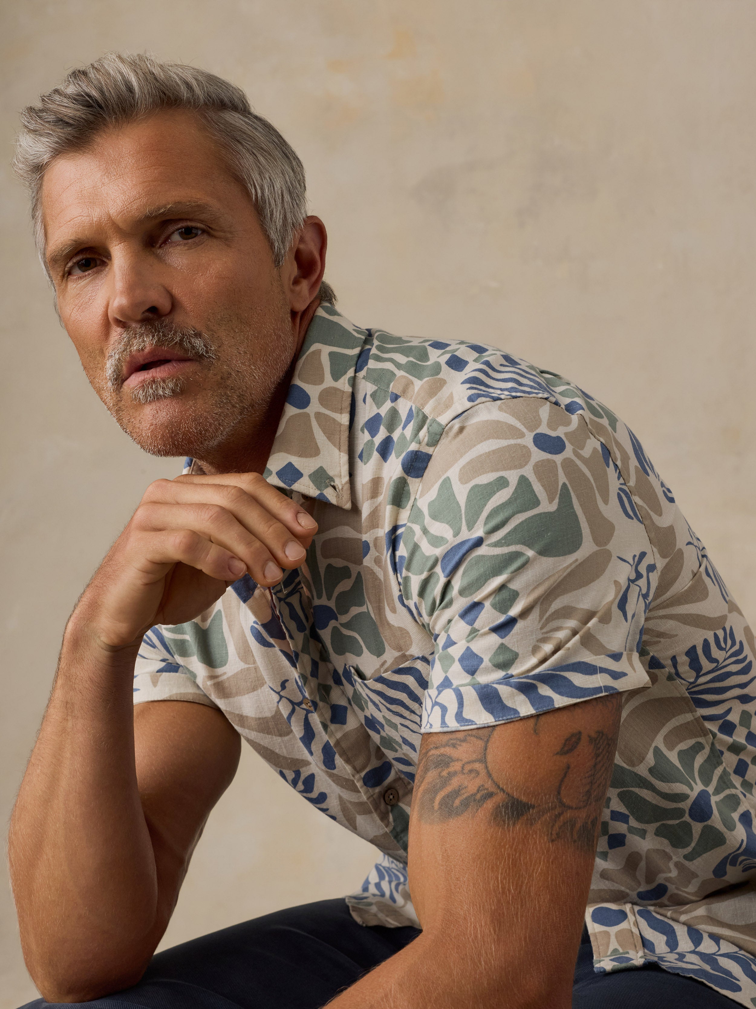 A man with gray hair and a mustache wearing a short-sleeve button-up shirt with a blue, green, and beige botanical pattern, posing against a textured beige wall.