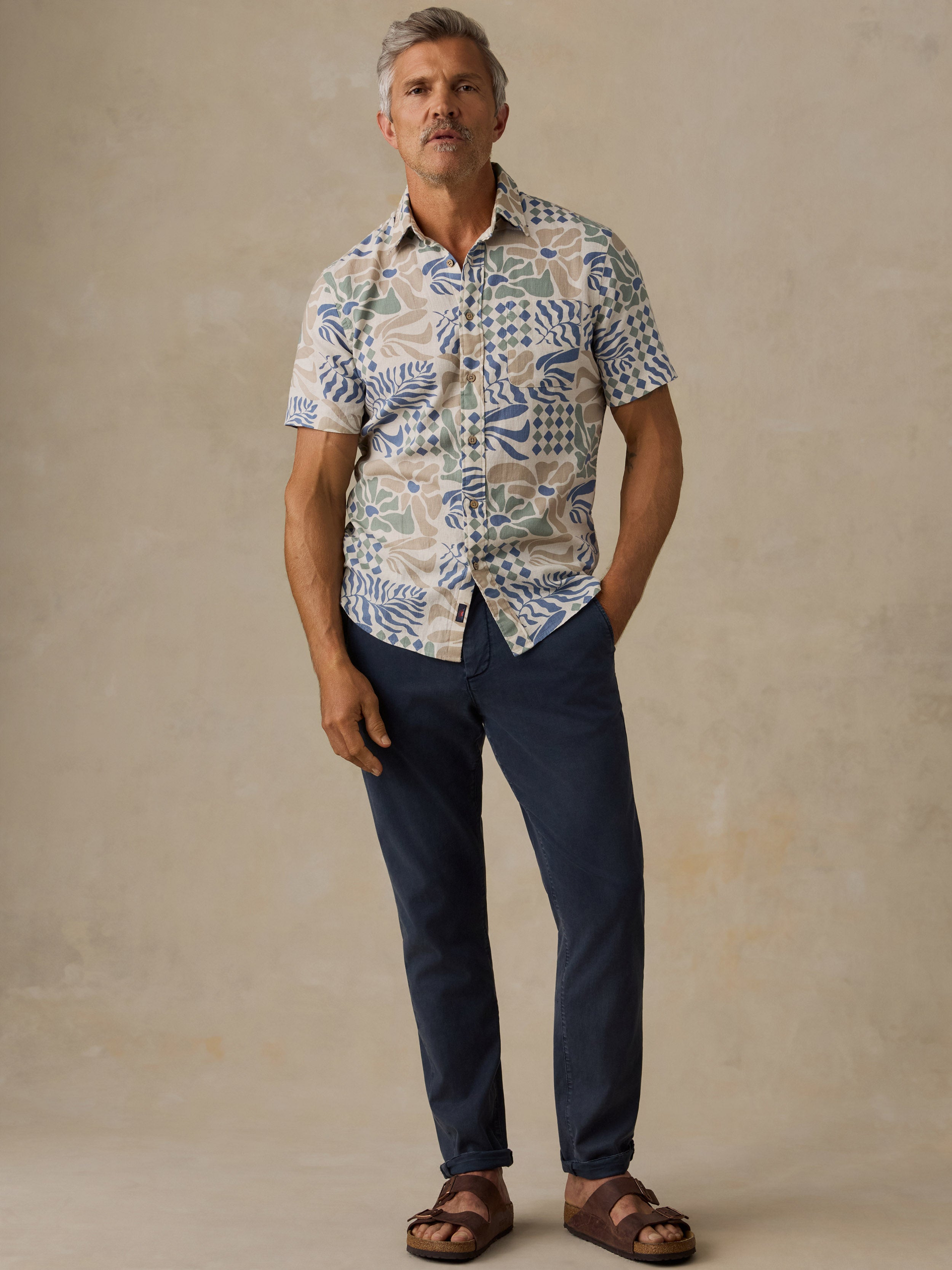 A man wearing a short-sleeve button-up shirt with a blue, green, and beige abstract pattern, paired with navy blue pants and brown sandals, standing against a textured beige studio backdrop.