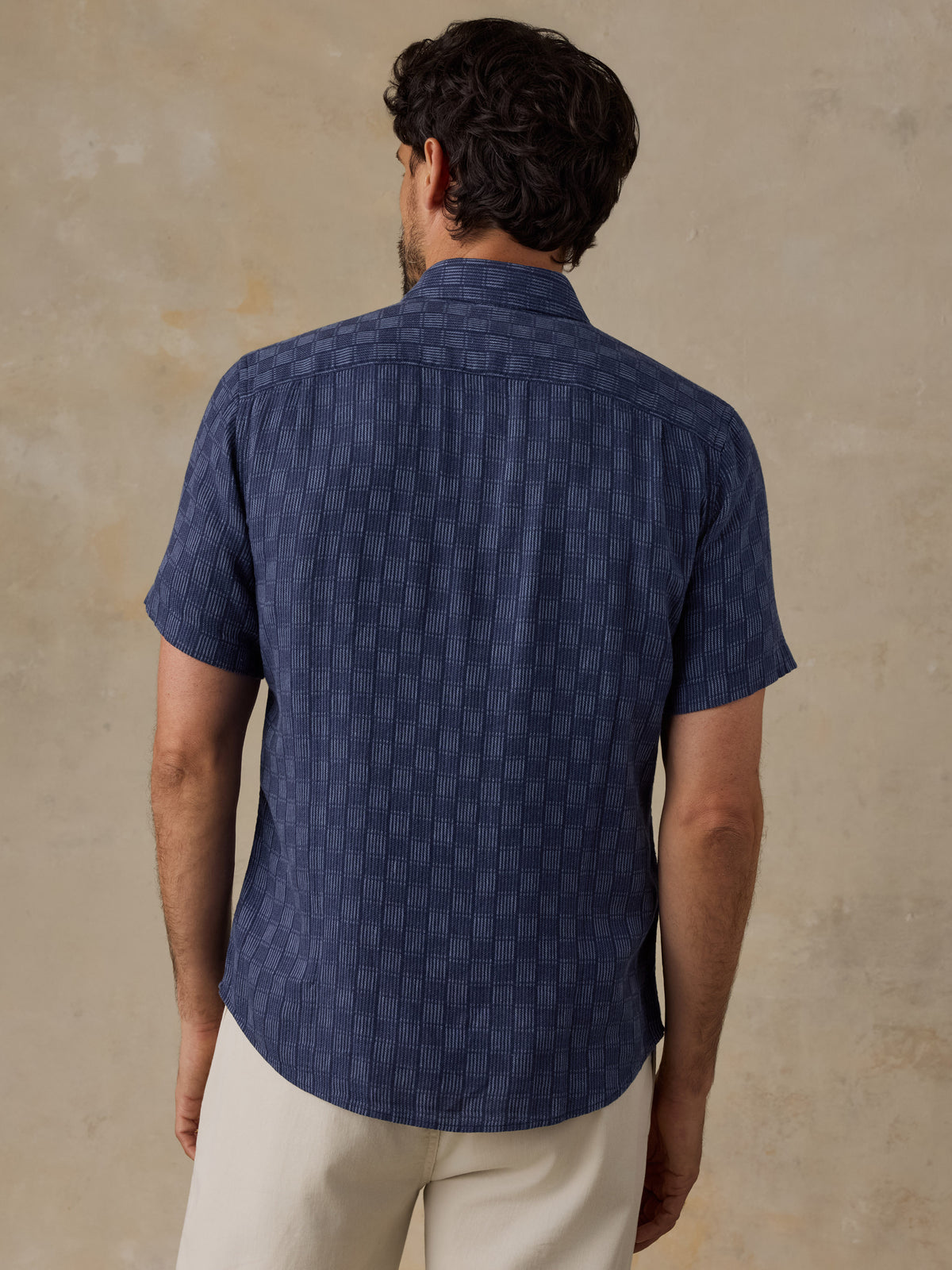 A man viewed from the back wearing a short-sleeve navy blue button-up shirt with a subtle vertical textured pattern, paired with light beige trousers, set against a textured beige wall., image 3 of 6