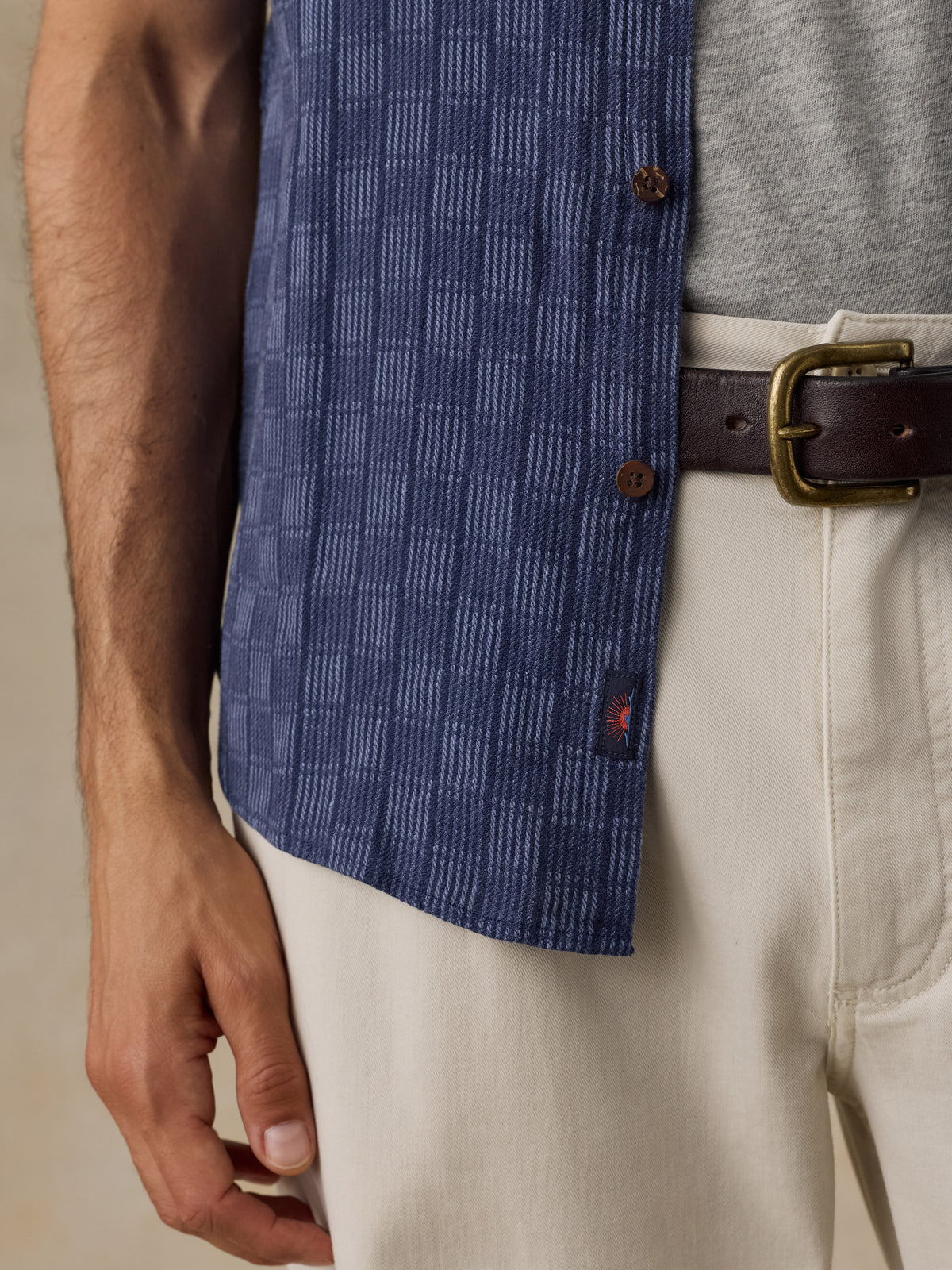 Close-up of a man wearing a blue patterned overshirt over a gray t-shirt, cream trousers, and a brown leather belt with a gold buckle., image 4 of 6