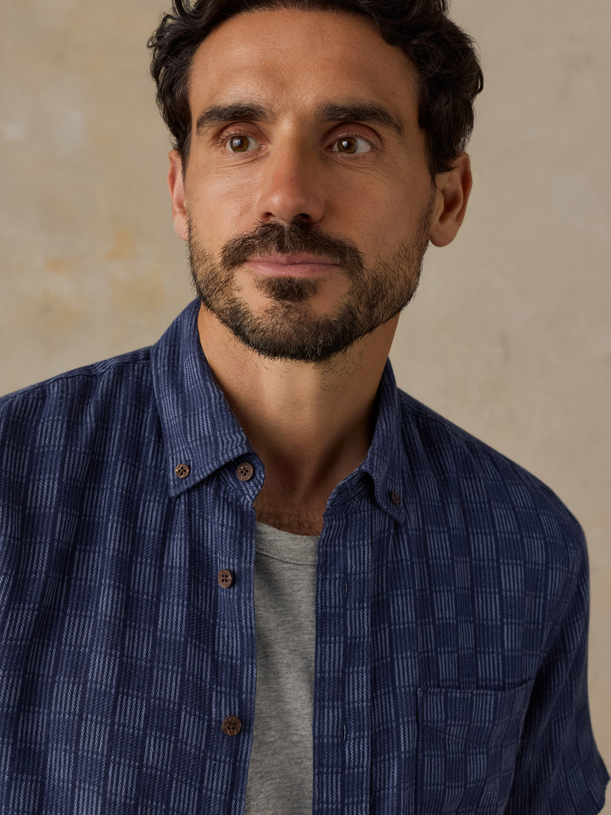 A close-up portrait of a man wearing a textured navy blue button-down shirt over a light gray t-shirt, set against a neutral beige background., image 2 of 6